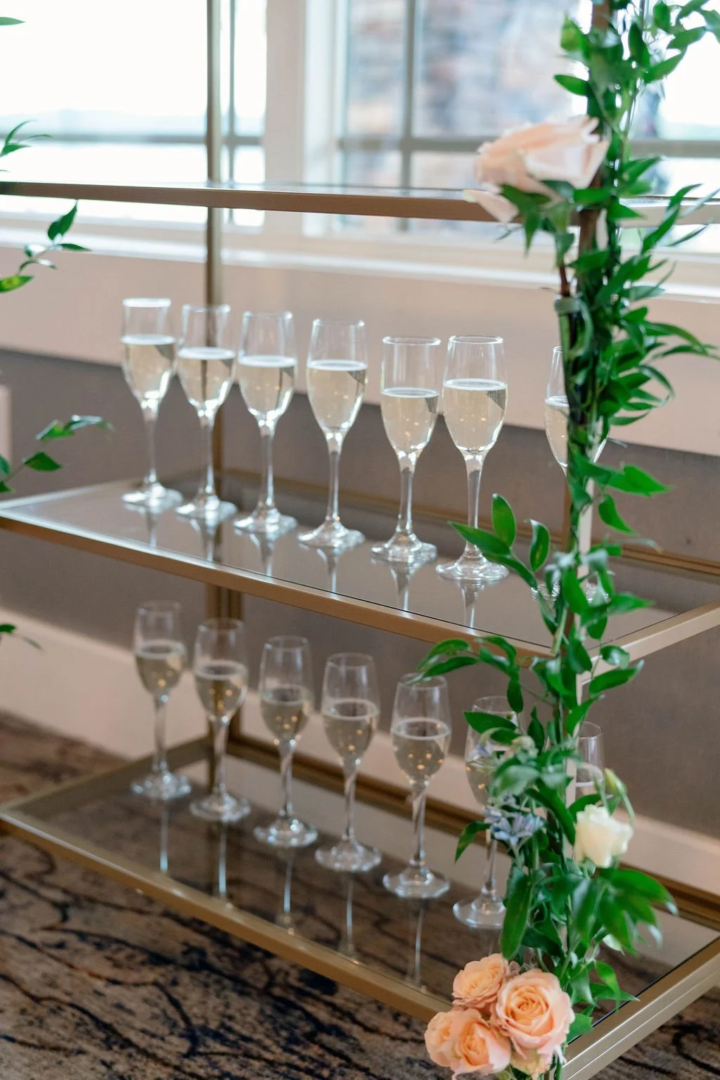 A three-tiered wedding display with champagne flutes filled with sparkling wine, decorated with pink roses and green foliage on the side, set on a metal stand near large windows.