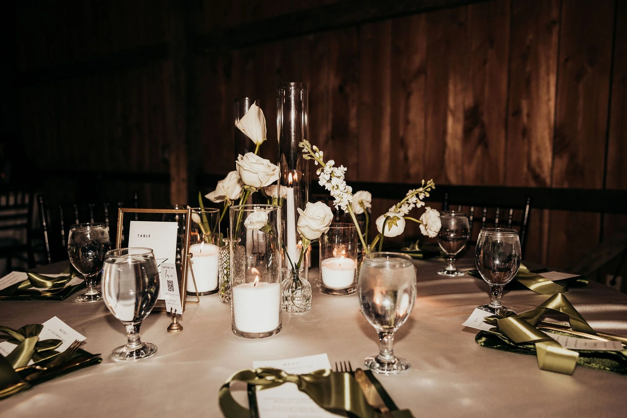 A decorated dining table with white roses and candles, set for an event or celebration in a dimly lit room with wooden walls.