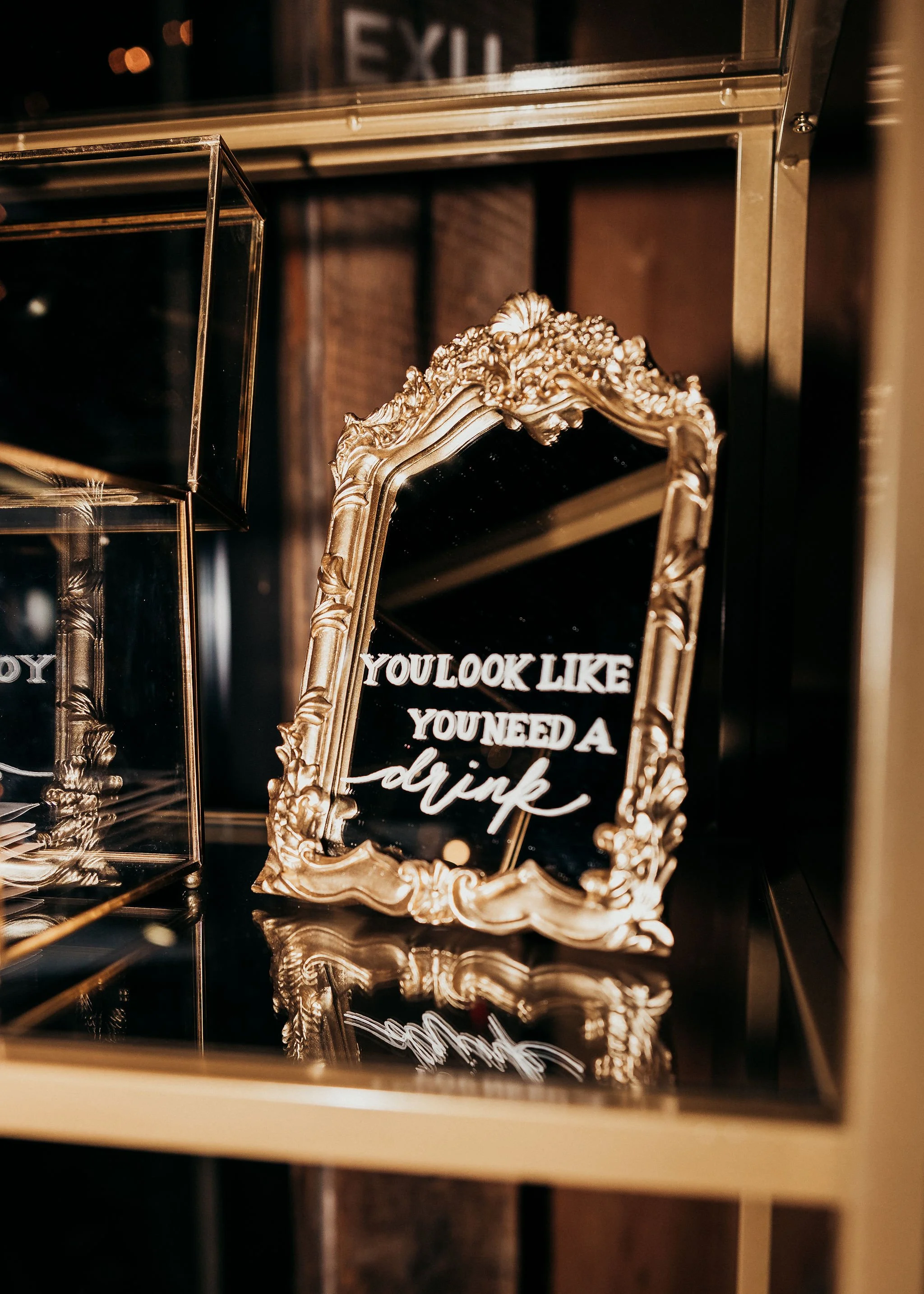 Decorative gold-framed mirror with a quote on a black background that reads, "You look like you need a drink." The mirror leans against a wall with a dark background, surrounded by glass display cases and metallic shelves.