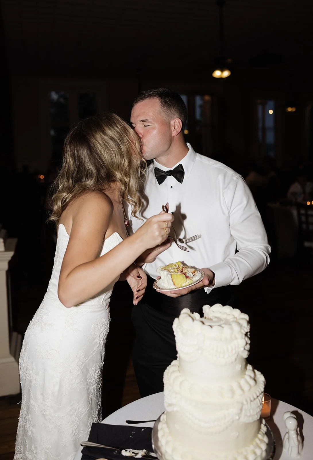 A bride and groom share a kiss at their wedding reception, with the bride wearing a white lace wedding dress and the groom in a white shirt with a black bow tie, as they feed each other cake.