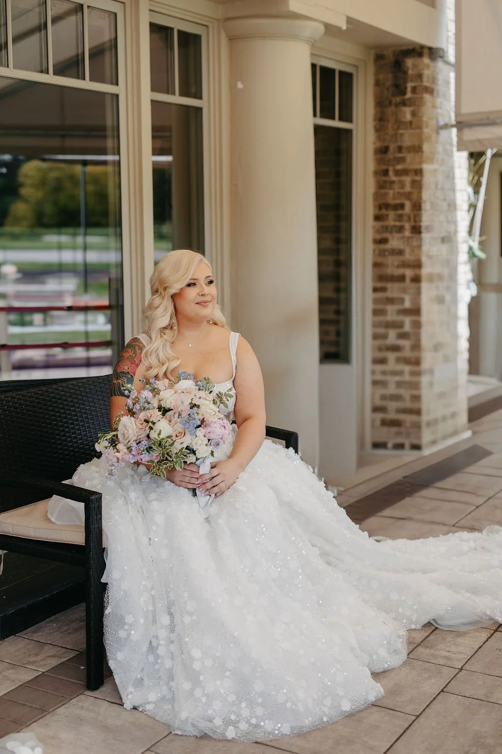 A bride with blonde hair in loose curls, wearing a white wedding gown with lace details, sitting on a black bench outdoors while holding a bouquet of pastel-colored flowers.