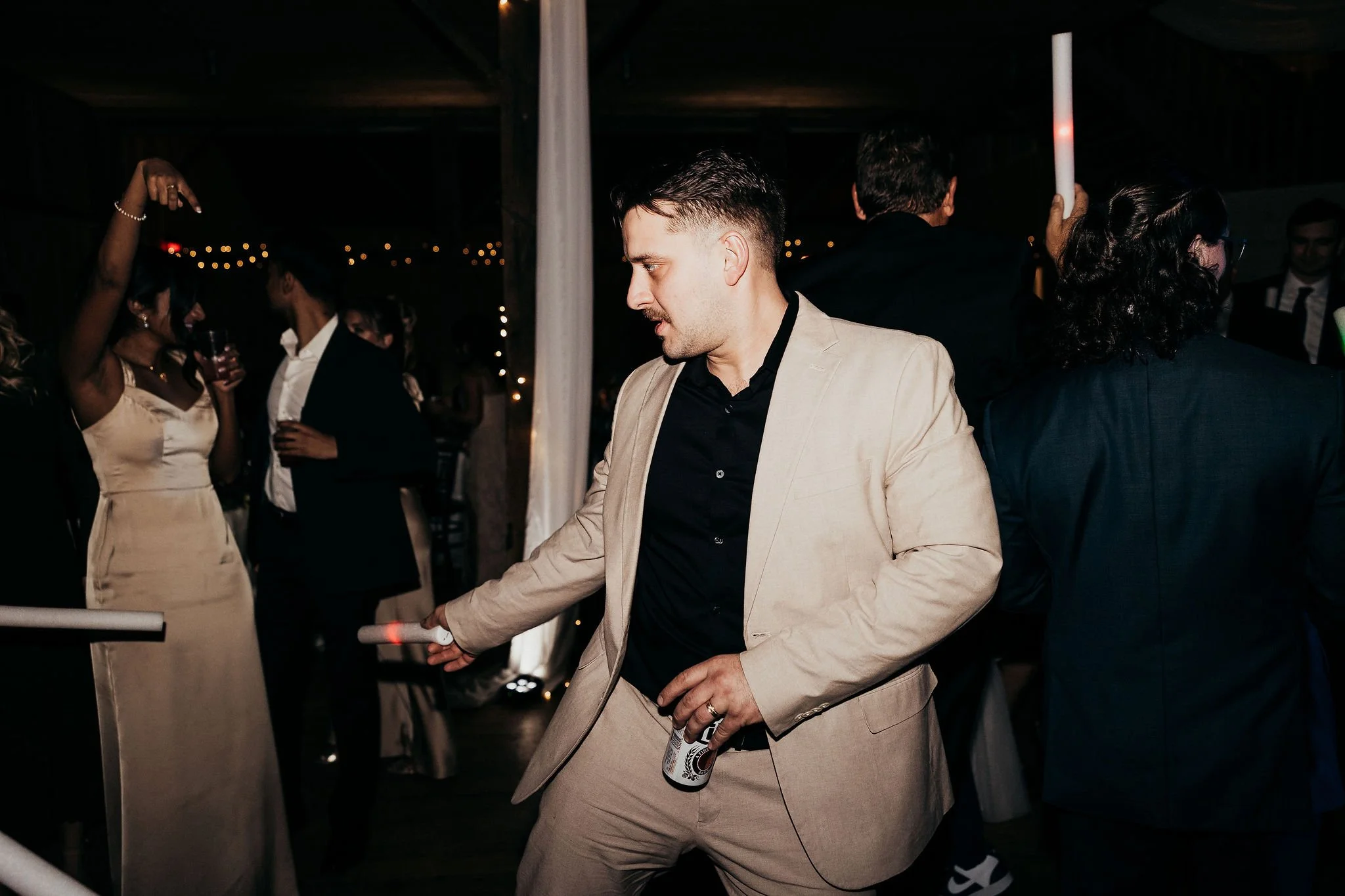 Man in beige suit dancing at a party with a beer in hand, surrounded by other guests in formal attire under dim lighting with string lights in the background.
