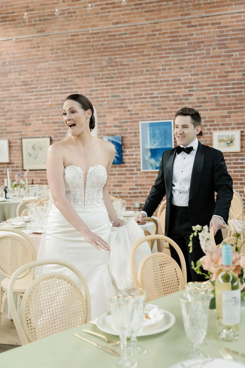 A bride and groom at their wedding reception, both smiling joyfully. The bride is in a strapless lace wedding dress, and the groom is in a black tuxedo with a bow tie. The setting is decorated with floral arrangements and elegant table settings.