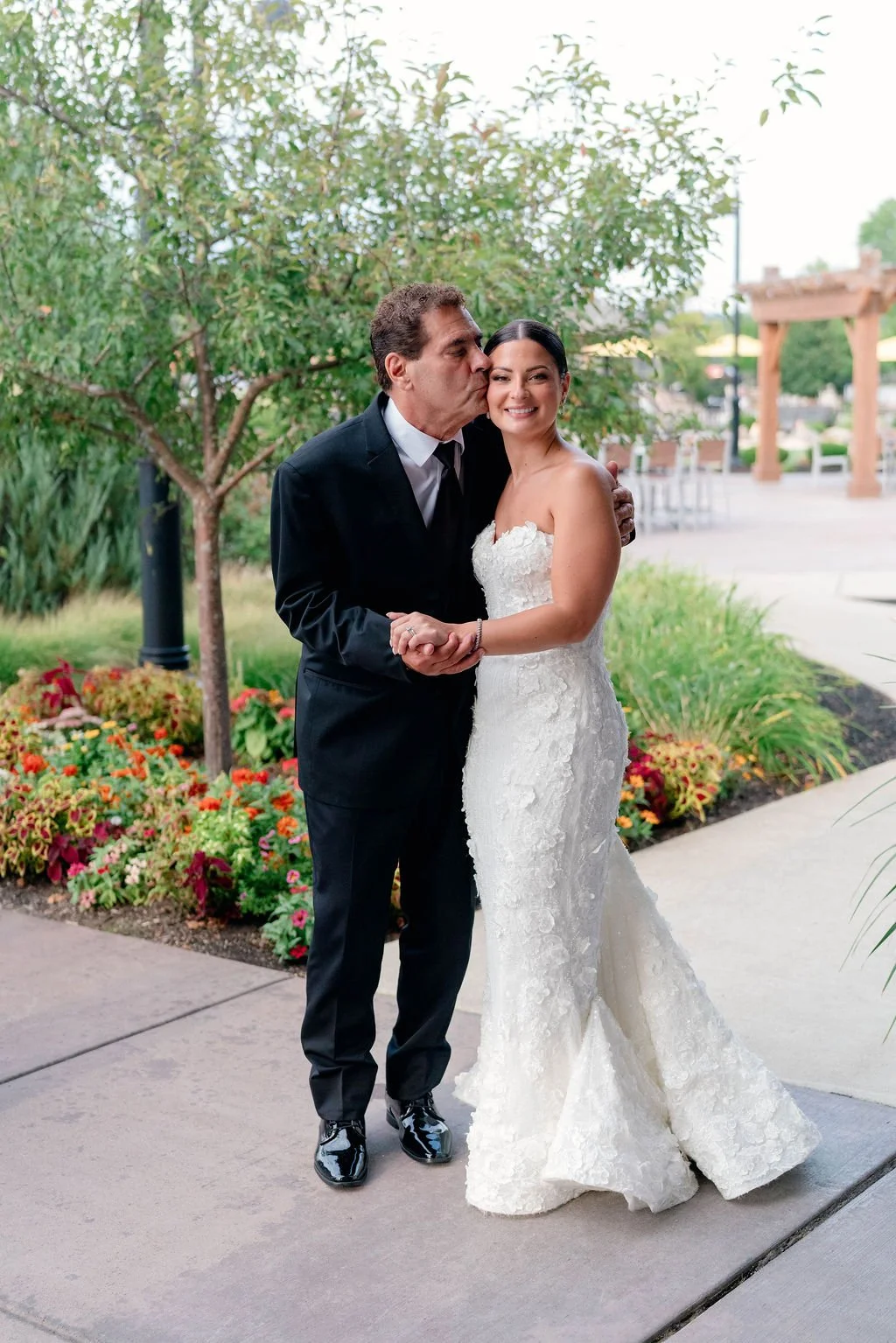 A bride and an older man in a tuxedo sharing a loving moment outdoors, with the man kissing the bride on the cheek. The bride is smiling in her strapless white wedding gown, standing on a garden path with flowers and greenery in the background.