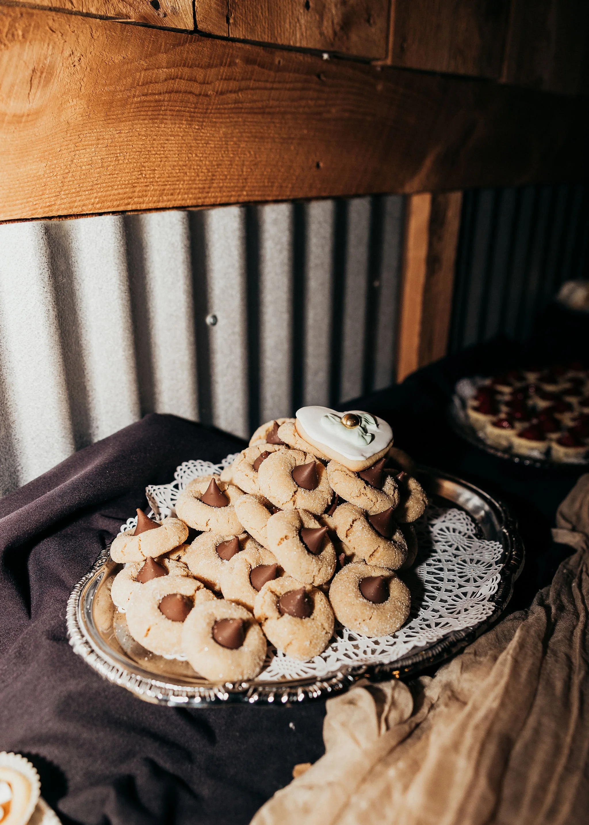 A silver tray filled with cookies topped with chocolate chips and a dollop of white icing, set on a dark cloth with a background of a rustic wooden wall and a corrugated metal panel.