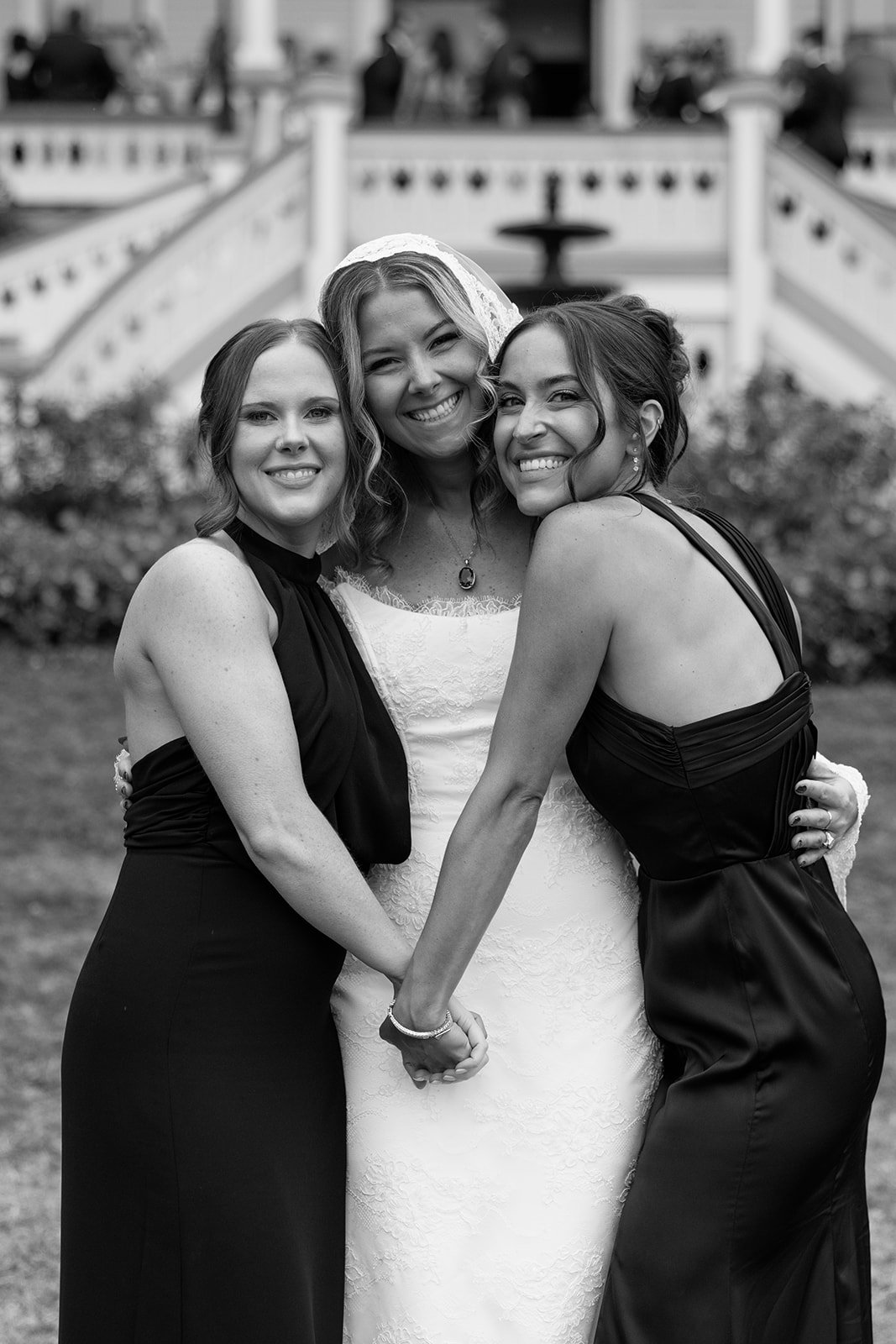 Three women, one in a wedding dress and two in black bridesmaid dresses, smiling and holding hands outdoors during a wedding celebration.