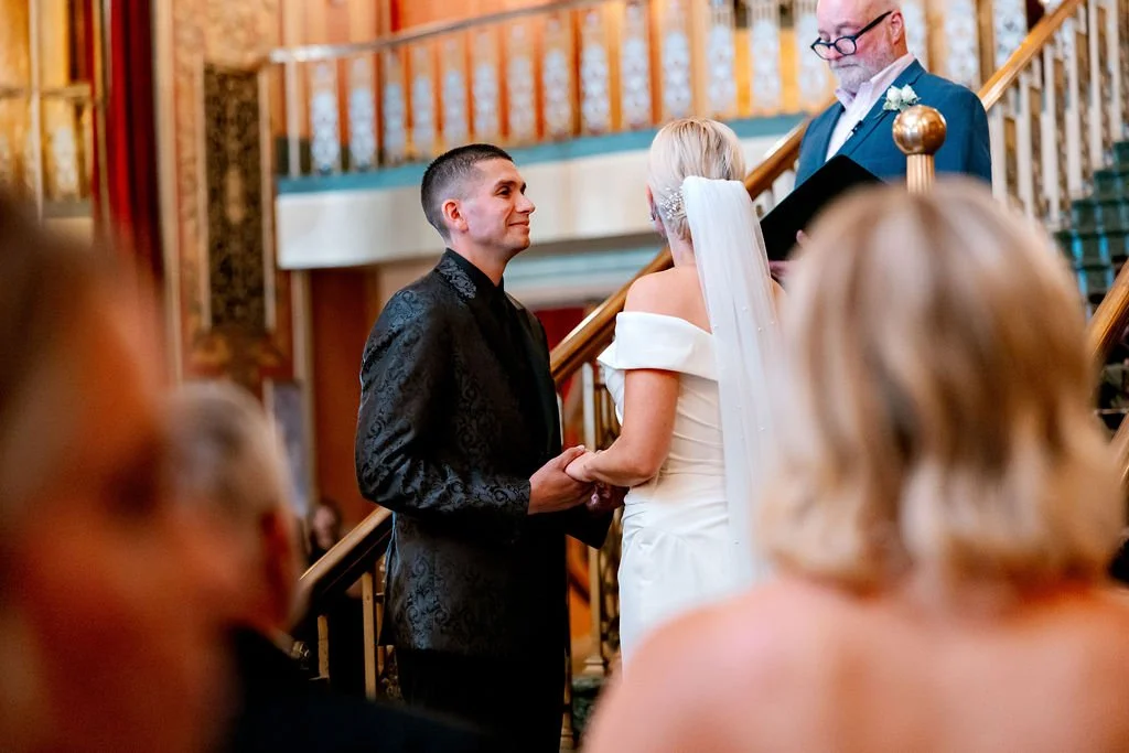 Bride and groom holding hands during wedding ceremony, officiant overseeing, indoor venue with staircase, guests in foreground.