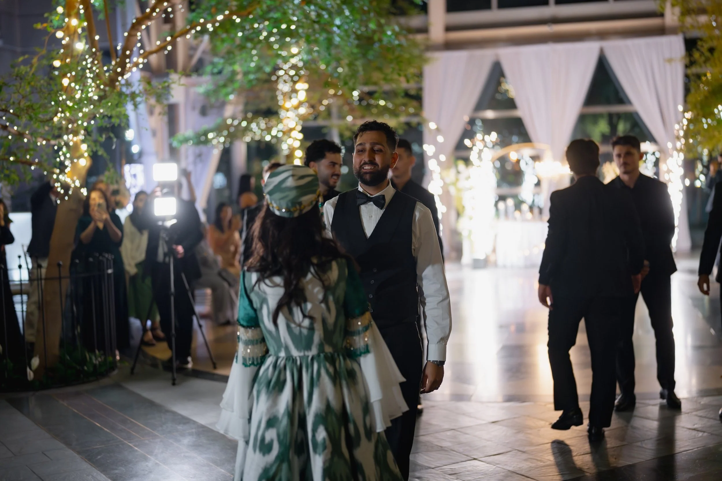 People dancing and socializing at an outdoor evening event decorated with string lights and trees.