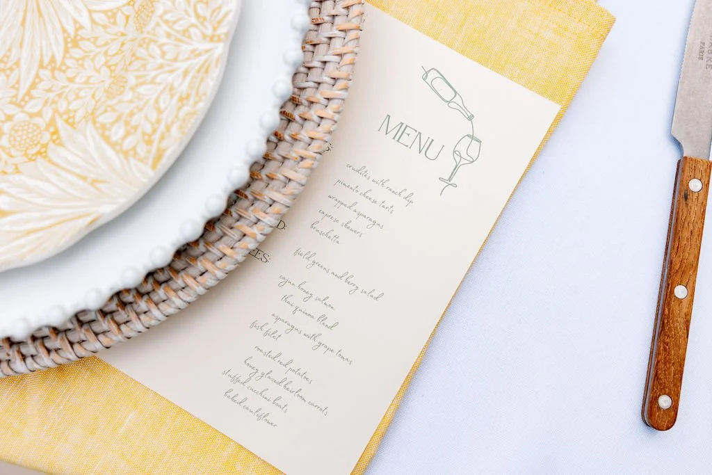 Partially visible decorative plates with a woven basket beneath them, a menu titled 'Menu' with a list of food items, a yellow napkin, a white tablecloth, and a wooden-handled knife.