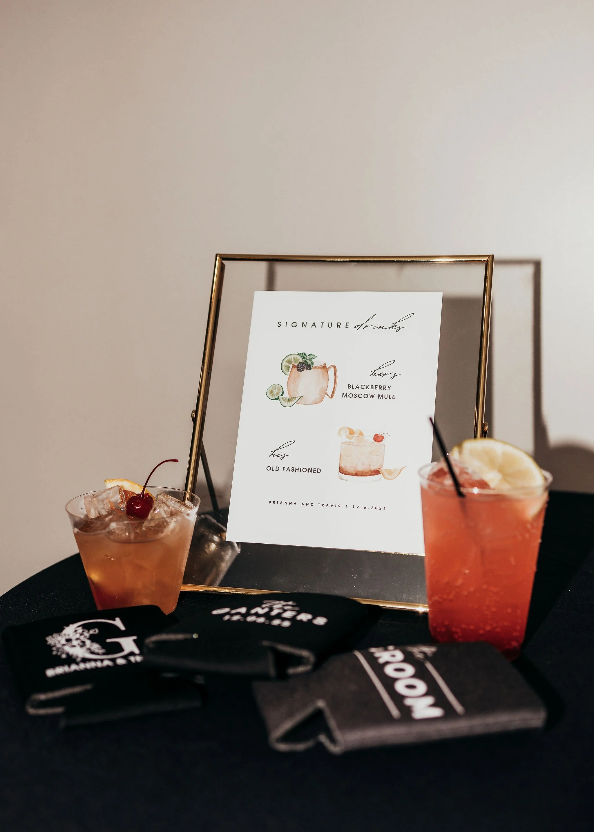 Two pink cocktails with ice and cherries on a black table, with a framed menu showing drinks like blackberry Moscow mule and old fashioned. The background is a plain, light-colored wall.