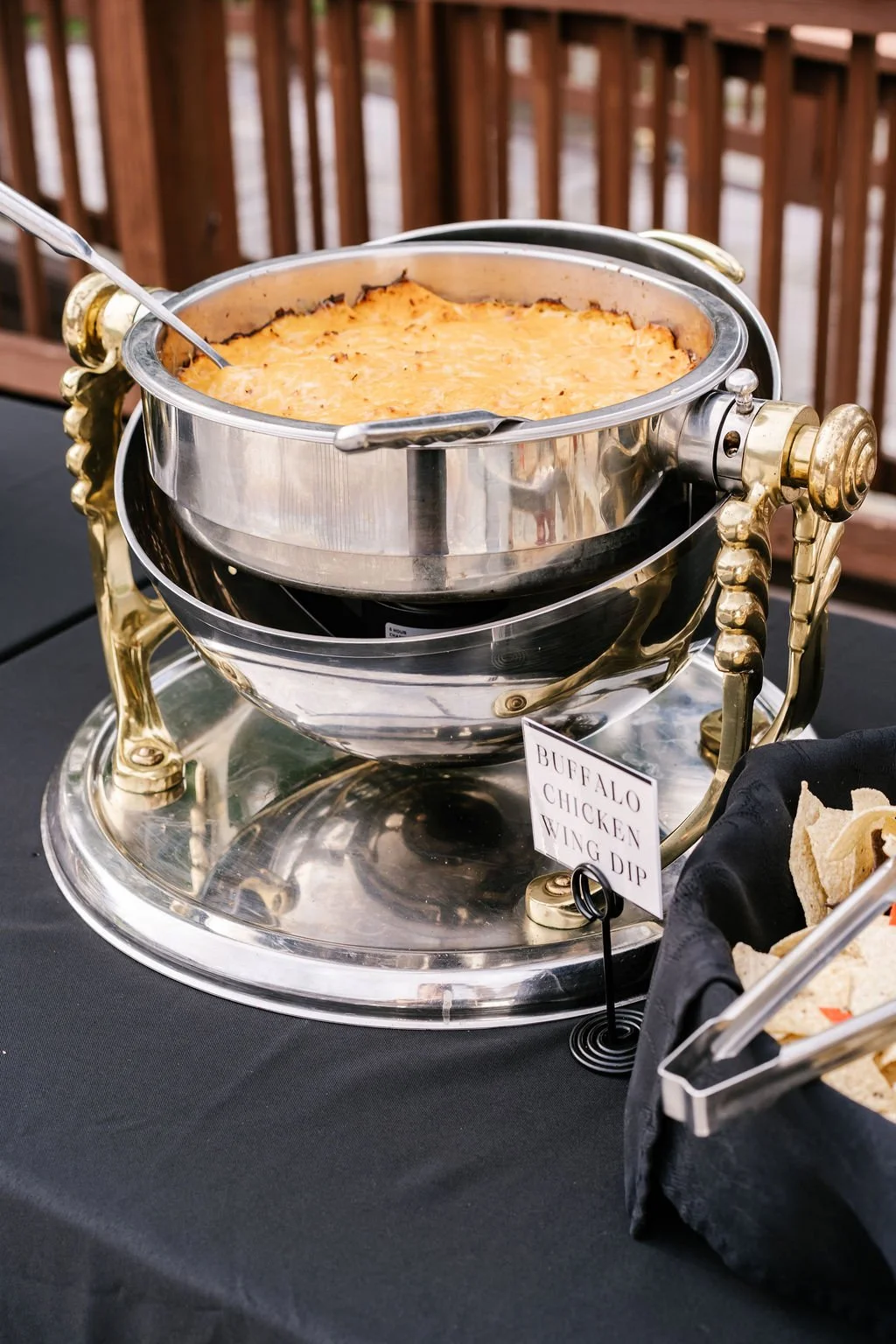 Chafing dish with Buffalo chicken wing dip on a black table, outdoors, with a wooden fence in the background.