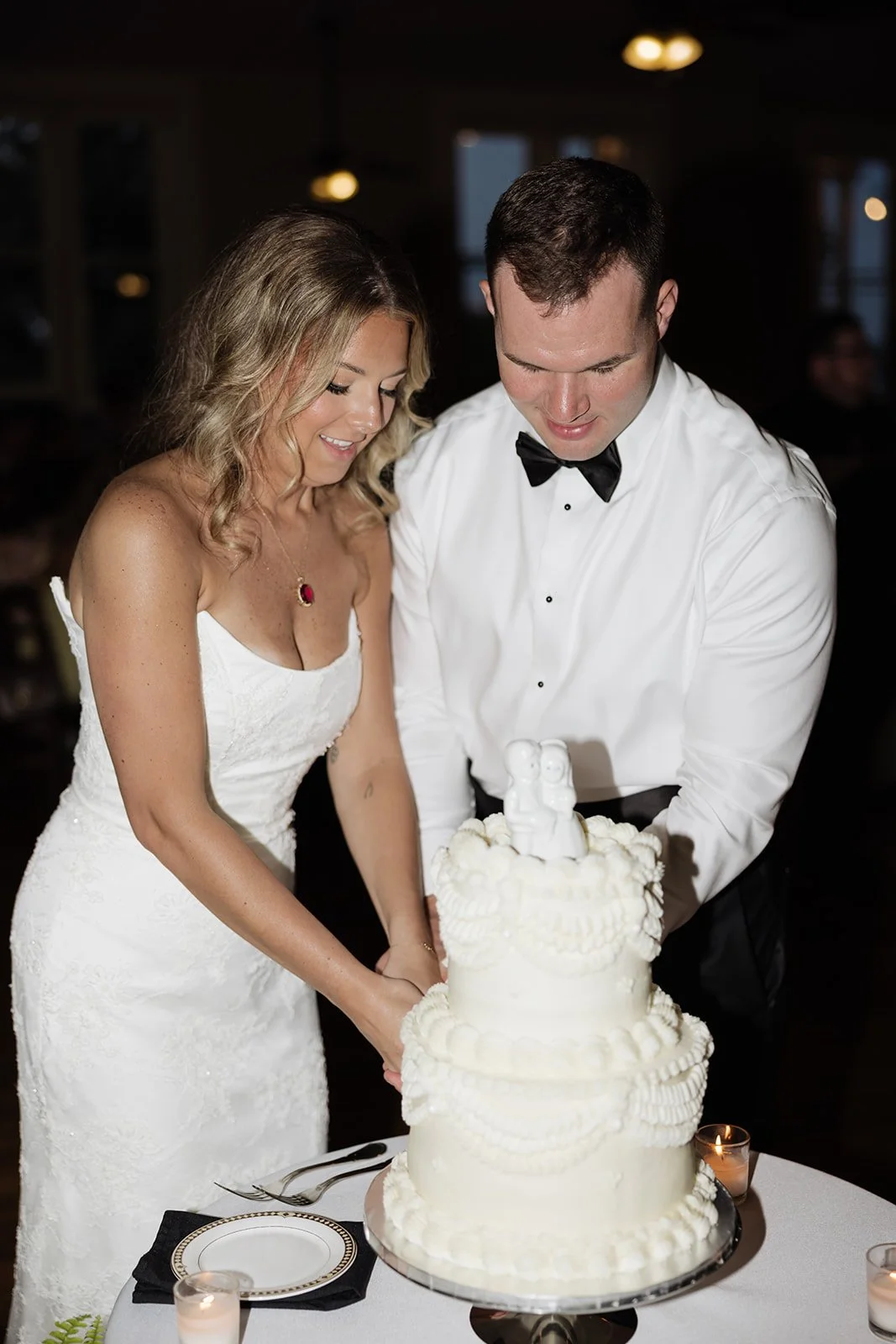 A newlywed couple cutting a white wedding cake with a bride in a white dress and a groom in a tuxedo, in a dimly lit reception hall.