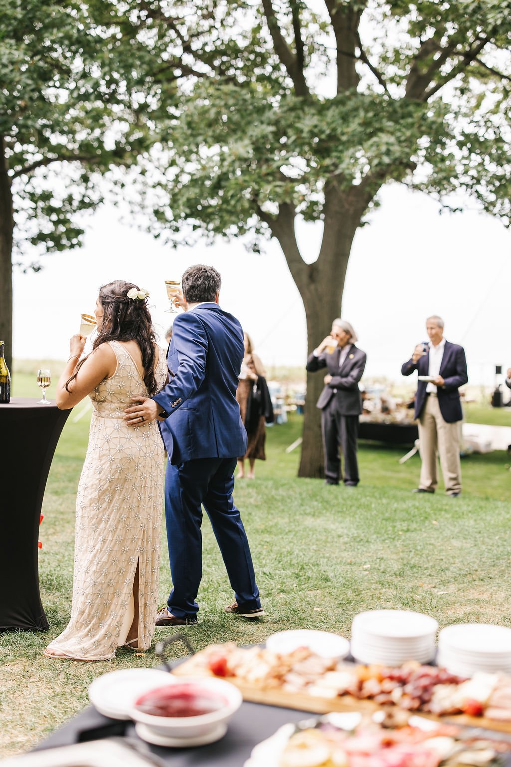 People at an outdoor celebration or wedding, standing around a table with drinks and food, under a large tree, with guests in formal attire in the background.