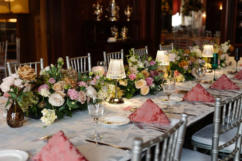 Elegant dining table set with pink and cream flowers, pink cloth napkins, and small lamps in a dimly lit, wood-paneled room.