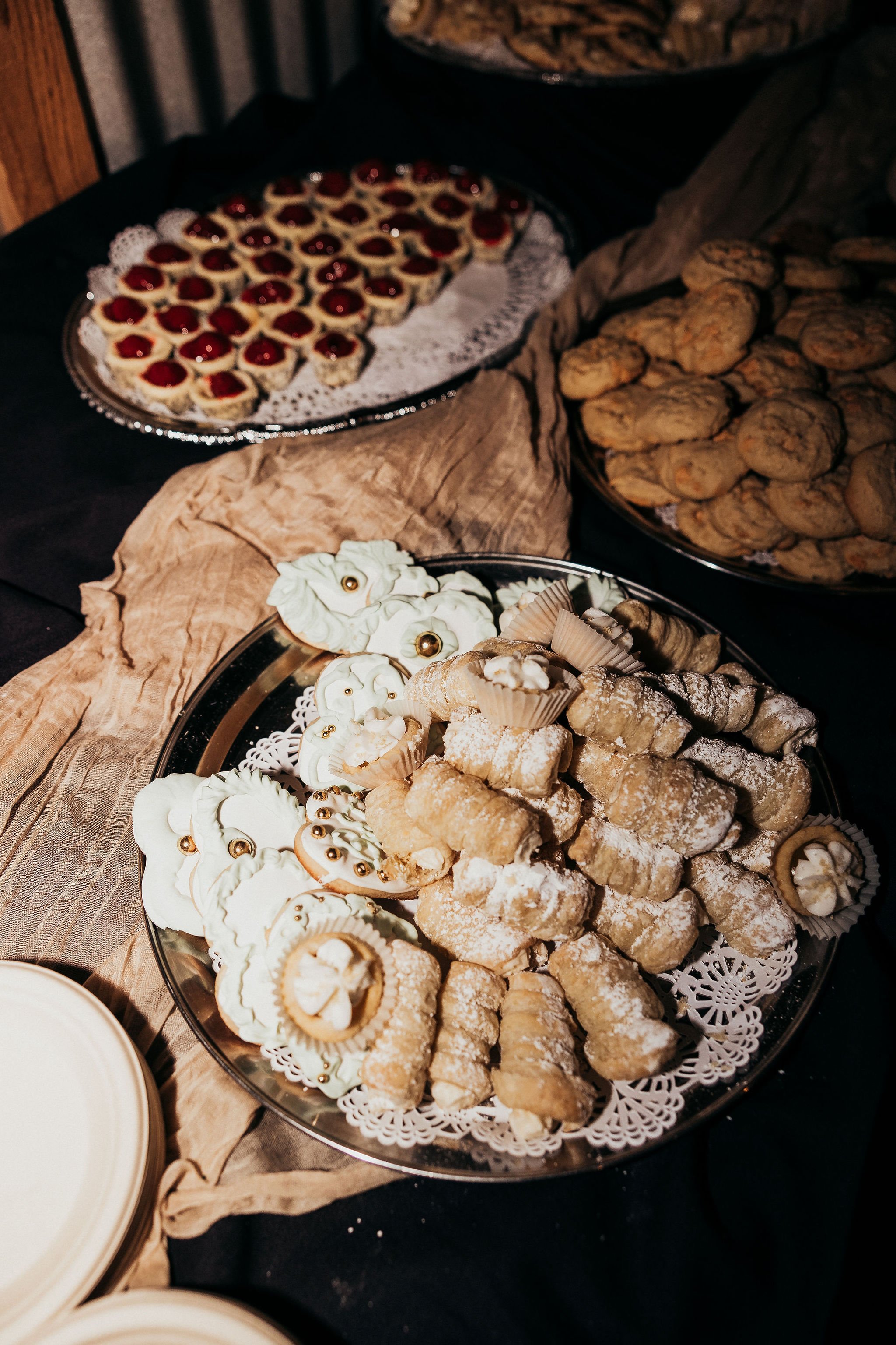 Tray of assorted cookies with decorative icing and gold accents, placed on a doily on a silver platter, with other trays of cookies in the background.