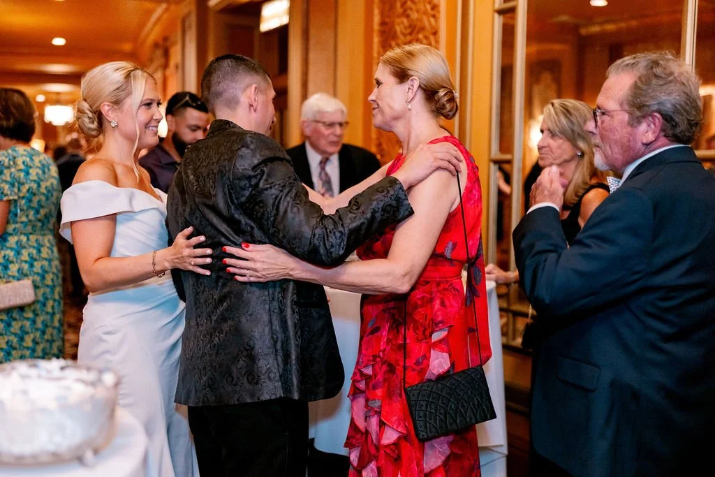 A group of elegantly dressed people at a celebration or wedding reception, dancing and socializing in a warmly decorated room.