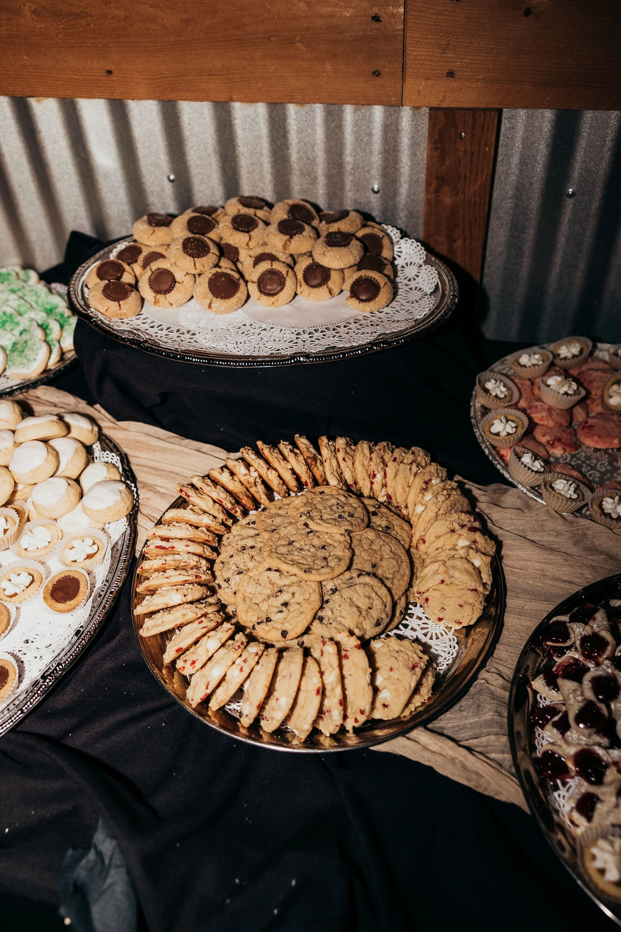Tray of assorted cookies including peanut butter cookies with chocolate centers, oatmeal cookies, and various decorated cookies on a table with a black tablecloth and a wooden backdrop.