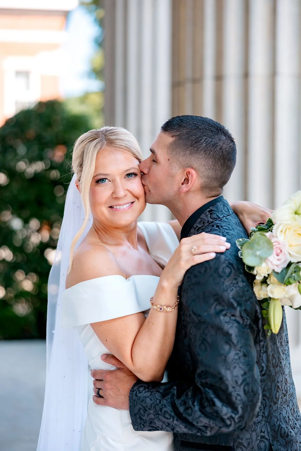 A bride with blonde hair and a white dress smiling as a groom kisses her on the cheek, holding a bouquet of flowers, standing outside in front of a building with columns.