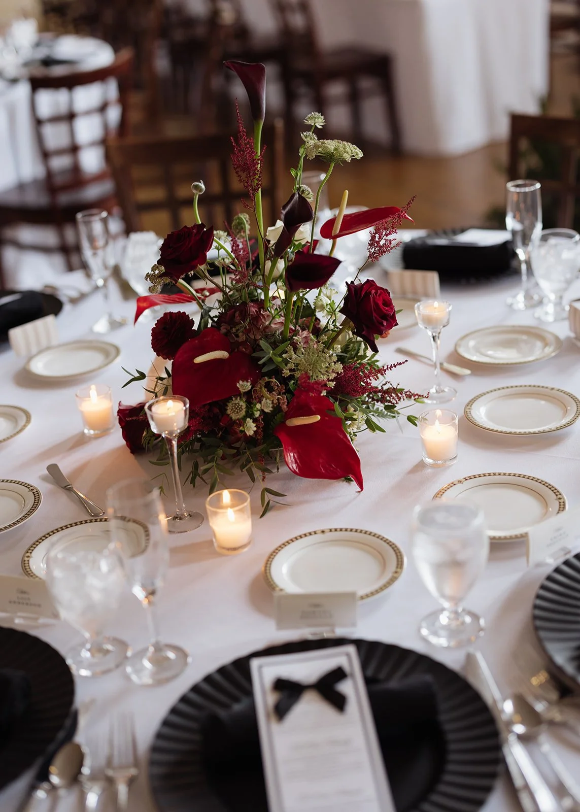 Elegant banquet table decorated with a large centerpiece of dark red and green flowers, surrounded by lit candles and set with white plates and glassware, in a warmly lit room with wooden chairs in the background.