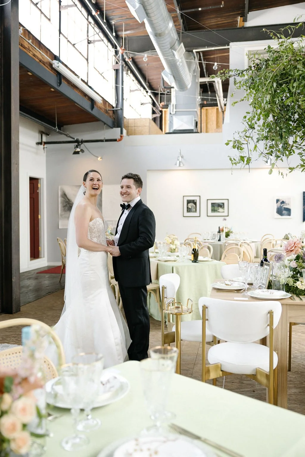 A bride and groom smiling and holding hands in a decorated reception hall with tables, chairs, flowers, and artwork on the walls.