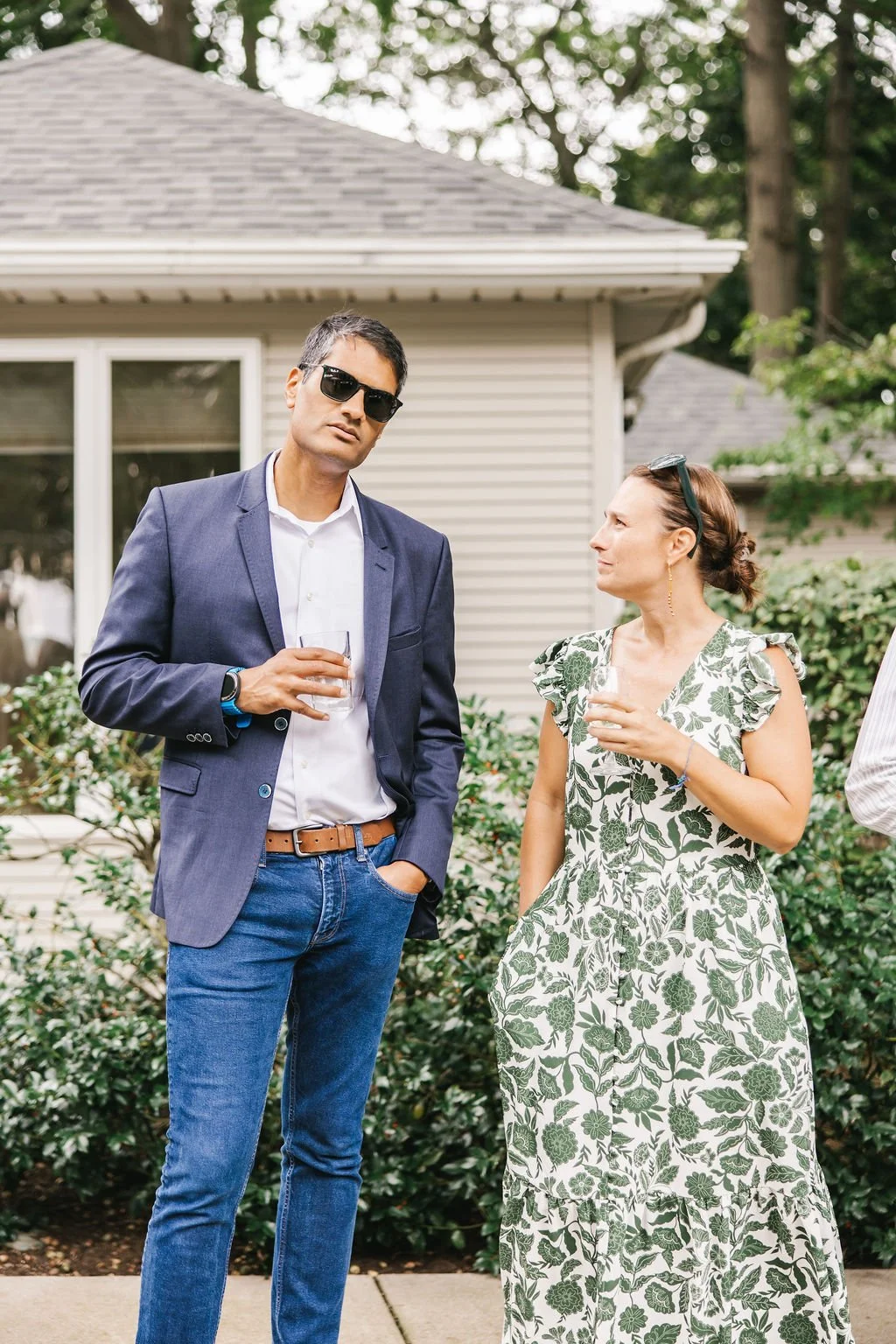 A man in a navy blazer, white shirt, jeans, sunglasses, and a watch, holding a drink, stands next to a woman in a white and green floral dress, holding a drink, and wearing sunglasses on her head, outdoors near a house and green bushes.