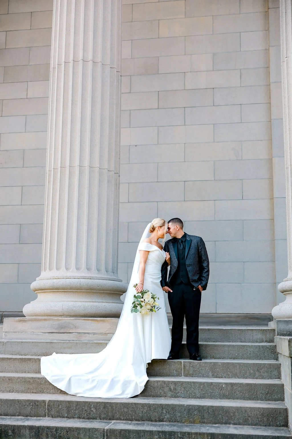 Bride and groom standing on steps in front of a large stone building with tall columns, leaning their foreheads together, with the bride holding a bouquet.