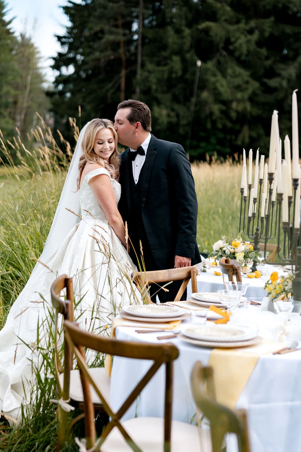 A bride and groom share a tender moment at an outdoor wedding reception in a grassy field with tall trees in the background. The bride wears a white wedding gown with a veil, and the groom is in a black tuxedo. The reception table is set with white p