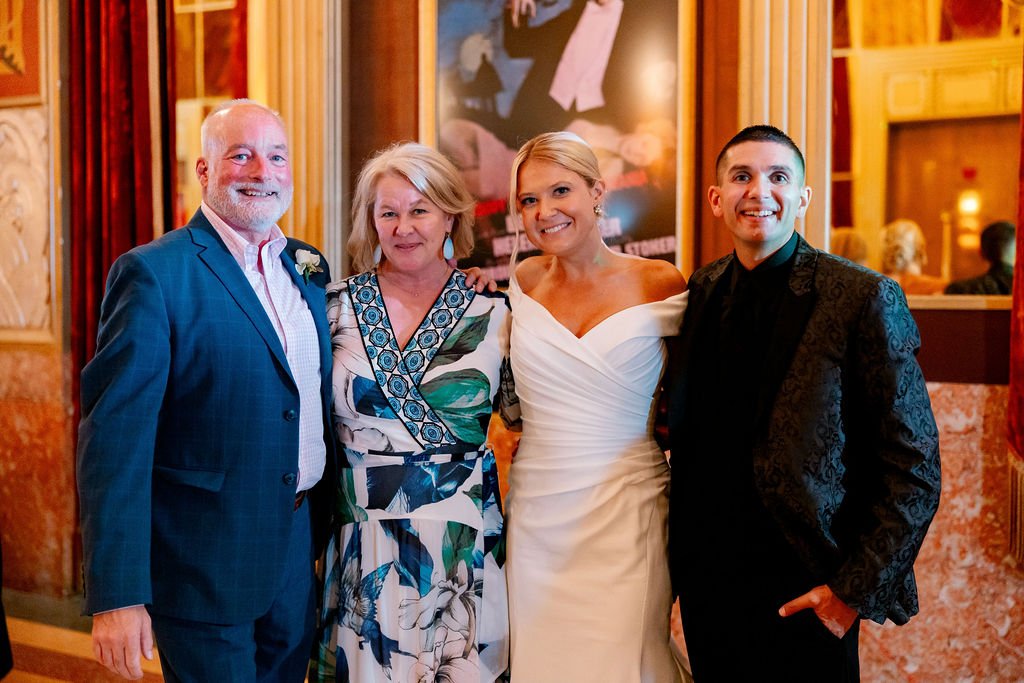 Group of four people at a wedding reception, with a bride in a white dress and groom in a black suit, standing between two older individuals, all smiling in a warmly decorated indoor venue.