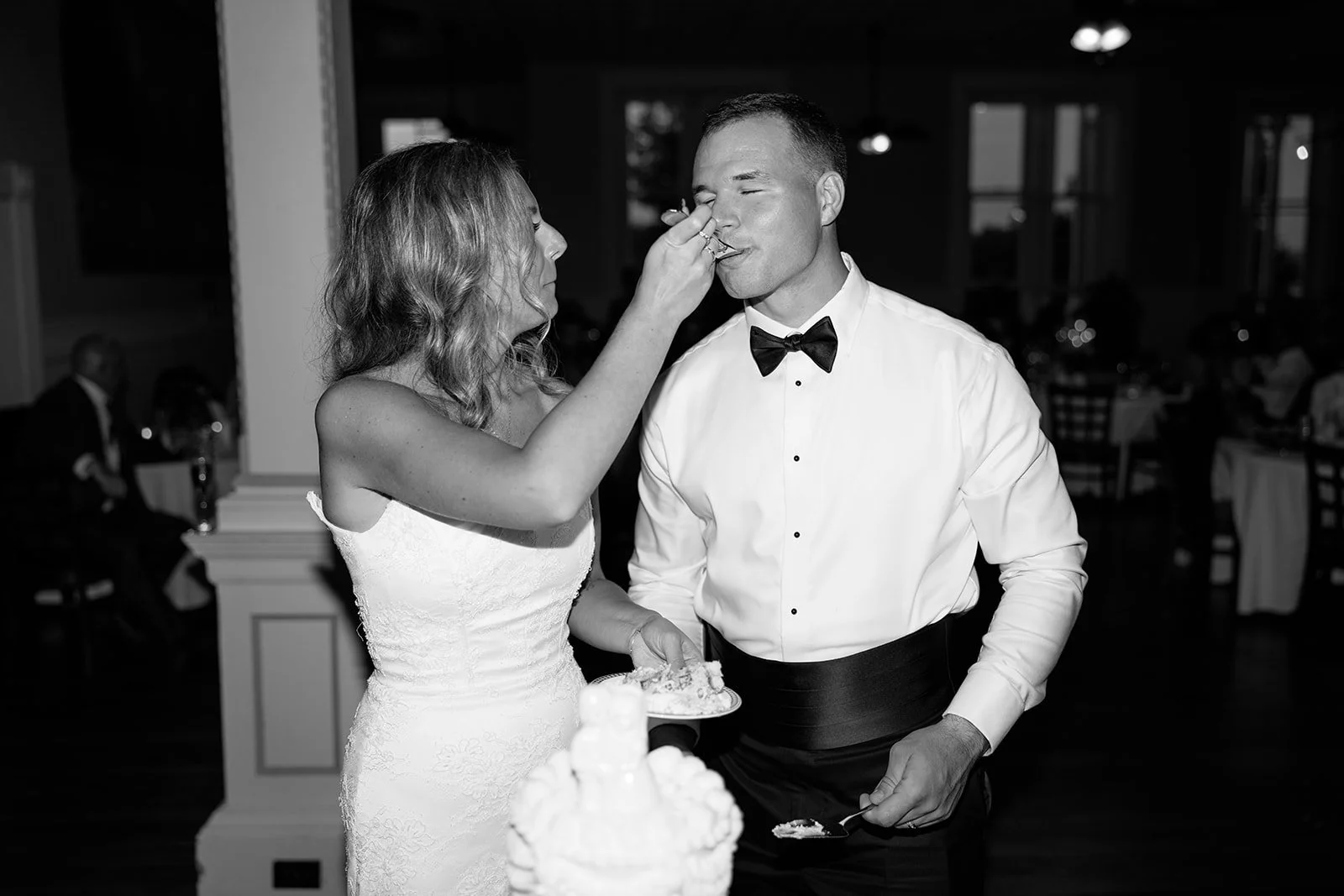 A bride feeds a slice of cake to a groom at a wedding reception, both dressed in formal attire indoors.