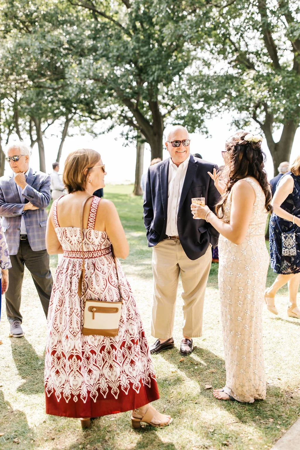 Group of people dressed in semi-formal attire outdoors, standing on grass with trees in the background, engaged in conversation at a social gathering or outdoor event.