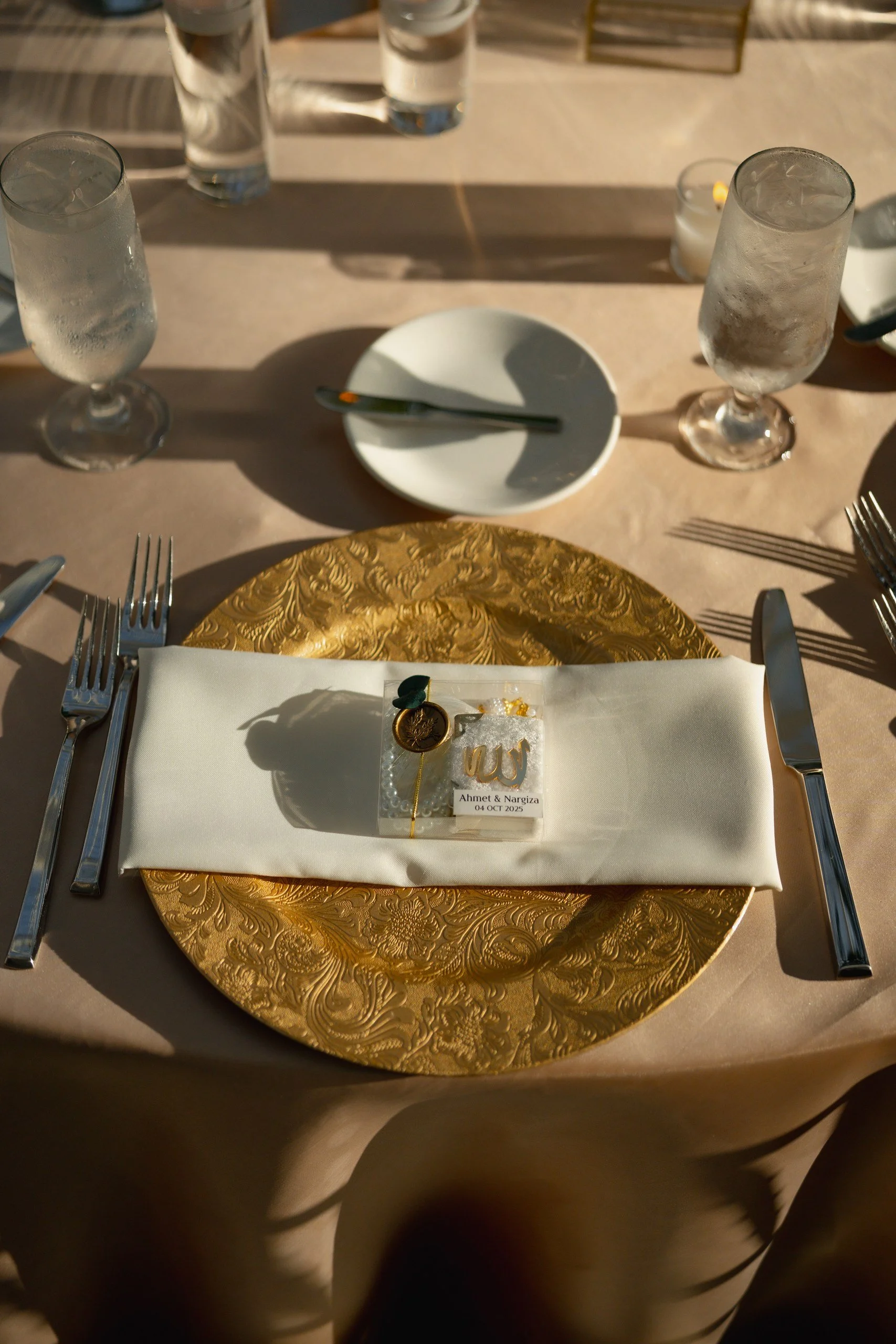 A table setting with a golden, ornate round charger plate, a white napkin, and silverware. On the napkin, there is a small clear box with a decorative gold and black item and a label that reads 'Ahmet & Nargiza, 04 OCT 2025.' Three champagne glasses 