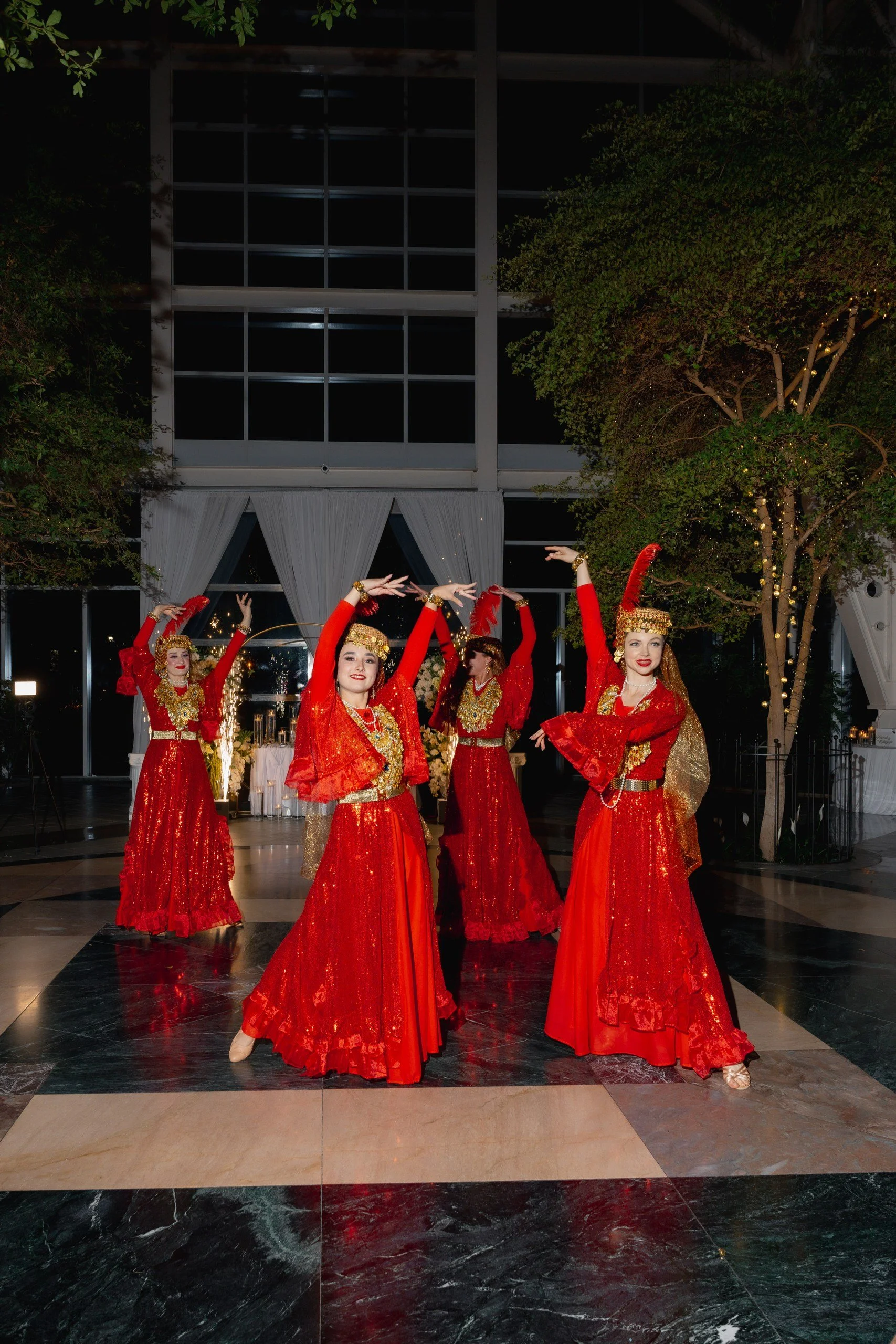 A group of women dressed in red traditional dresses with gold embellishments, performing a dance in an indoor venue with a polished black and white marble floor, surrounded by green trees and decorated with string lights.