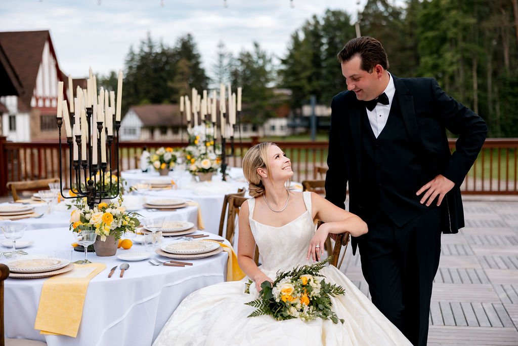 A bride and groom smiling at each other during their wedding reception outdoors, with decorated tables and a scenic background of trees and houses.