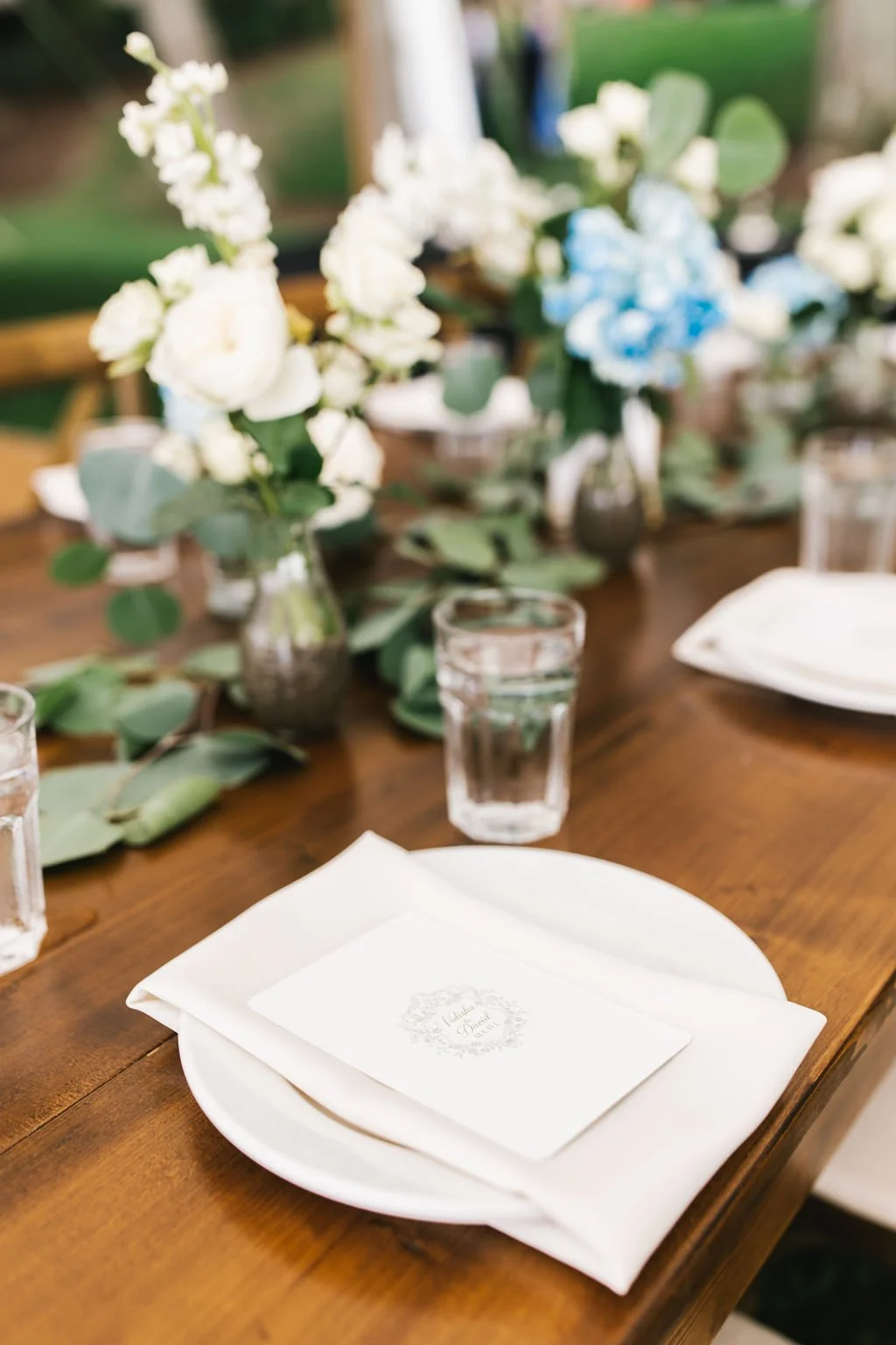 A close-up of a dining table setting with white plates, a white napkin, a glass of water, and a floral centerpiece with white and blue flowers in vases, set on a wooden table at an outdoor event.