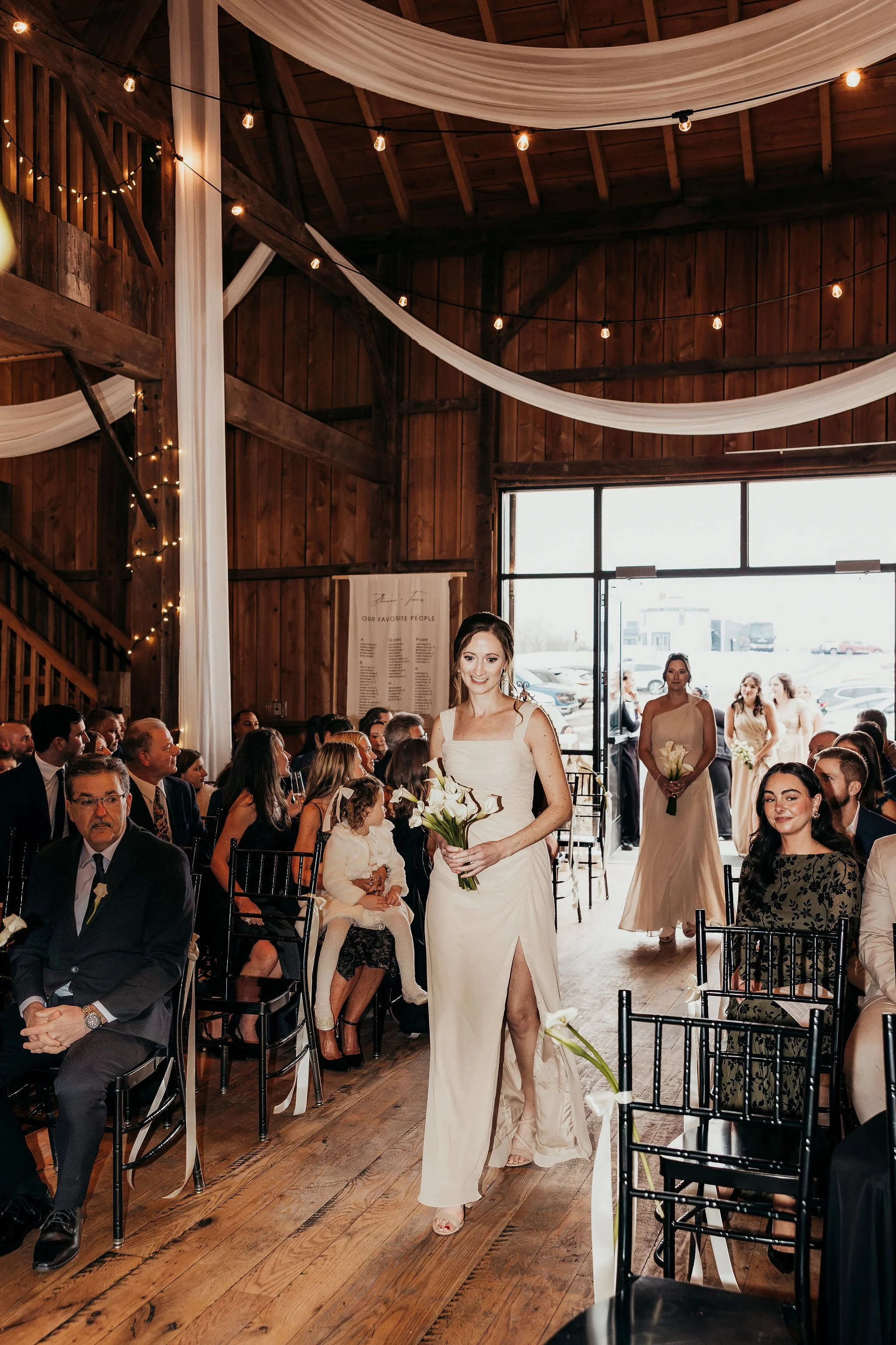 A bride walking down the aisle holding a bouquet of white flowers at a wedding ceremony inside a rustic wooden venue.