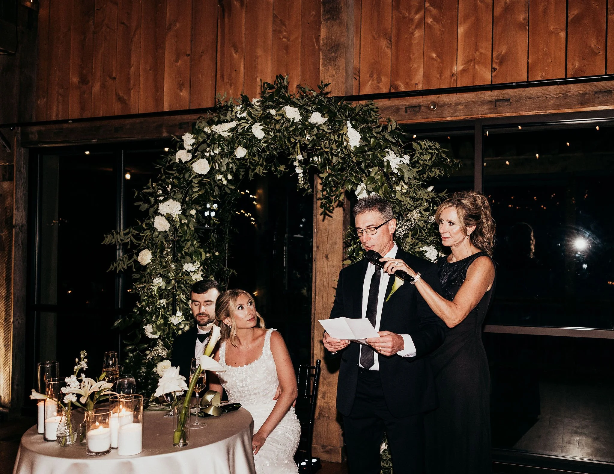 A wedding reception scene showing a man and woman reading vows or a speech, with the bride seated, surrounded by floral decorations and candles on the table, in a rustic wooden decorated venue.