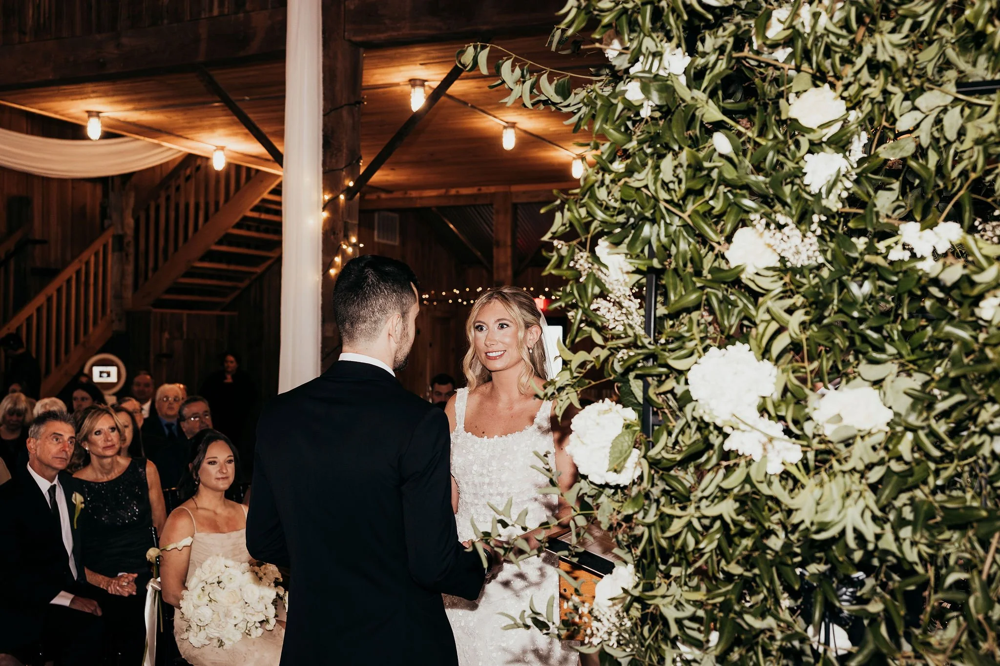 A bride and groom exchange vows at an indoor wedding ceremony, surrounded by guests seated in the background. The scene is decorated with lush greenery and white flowers, with warm lighting and wooden interior elements visible.