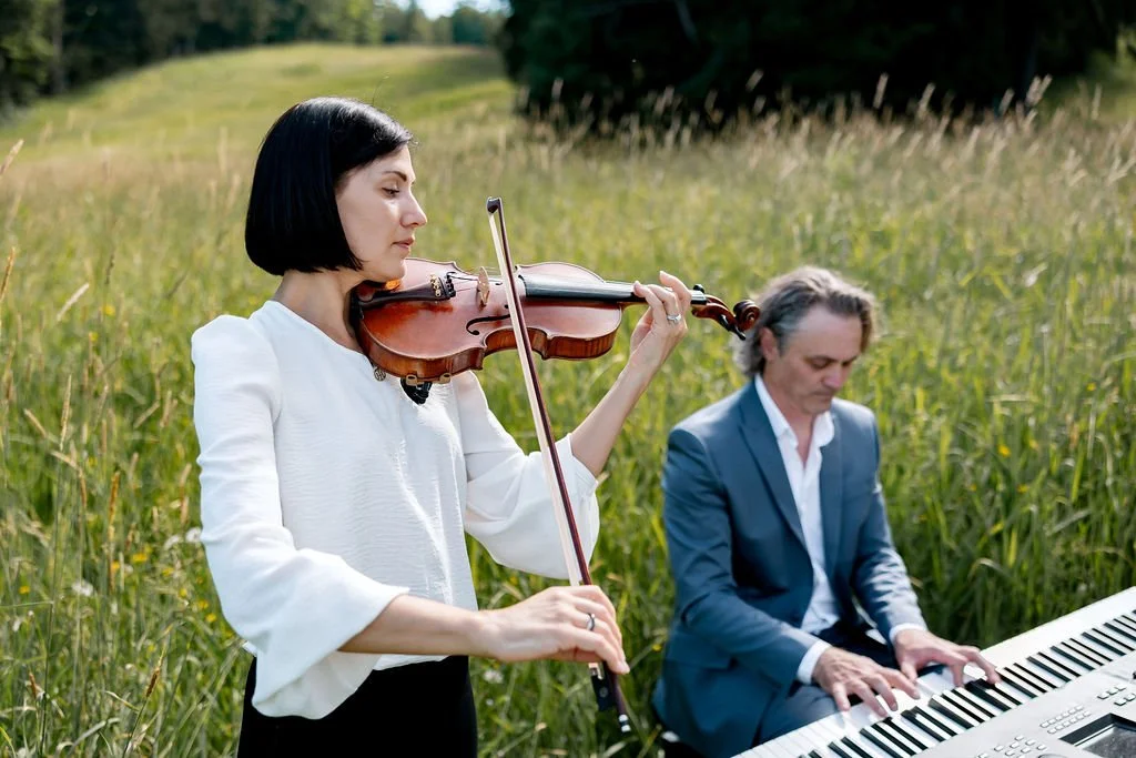 A woman playing a violin in a grassy field with a man playing a keyboard beside her.