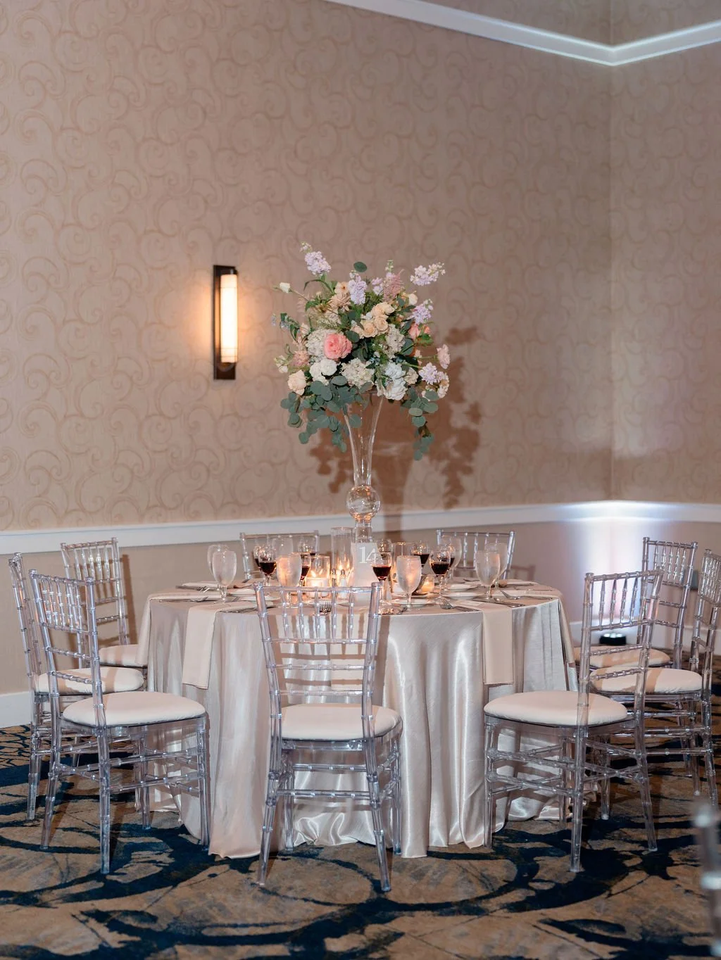 Elegant banquet table setup with a tall floral centerpiece, surrounded by transparent chairs, set with wine glasses and candles, in a softly lit ballroom with patterned wallpaper.