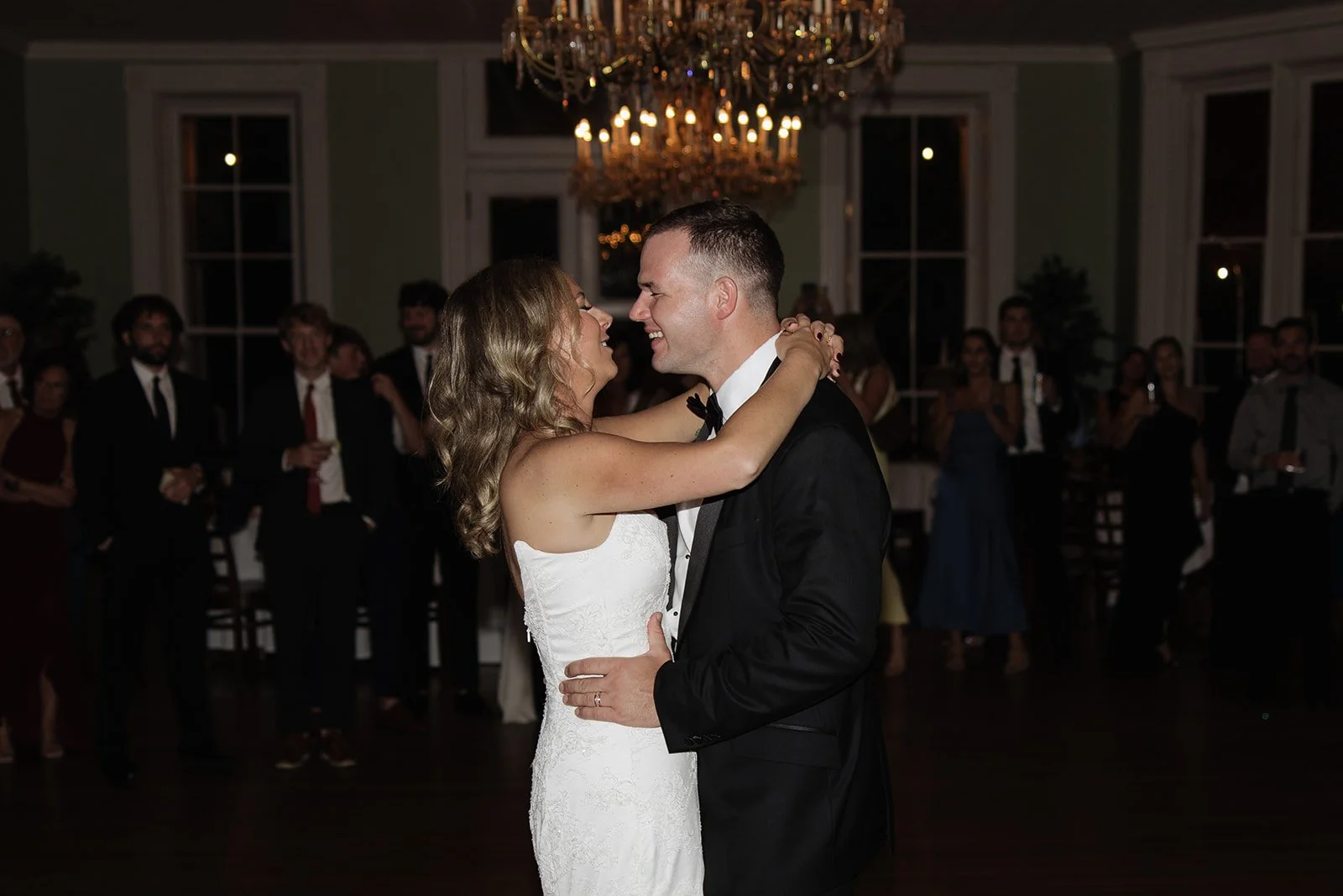 A bride and groom dance together at their wedding reception in a decorated ballroom, surrounded by guests in formal attire, with a chandelier hanging from the ceiling.