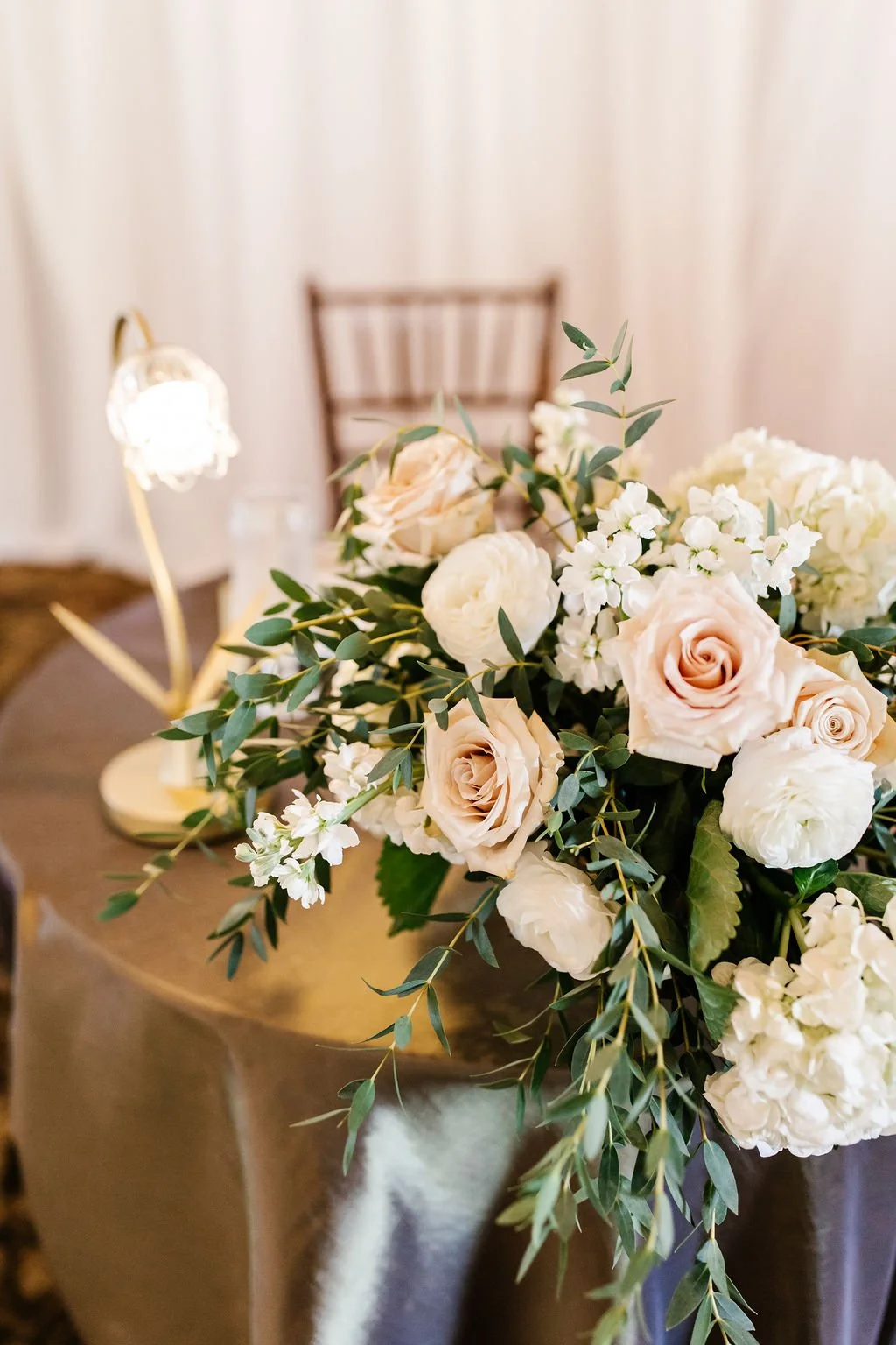 A floral centerpiece with white and blush roses, white hydrangeas, and green leaves on a brown table, with a blurred background of a chair and white curtain.