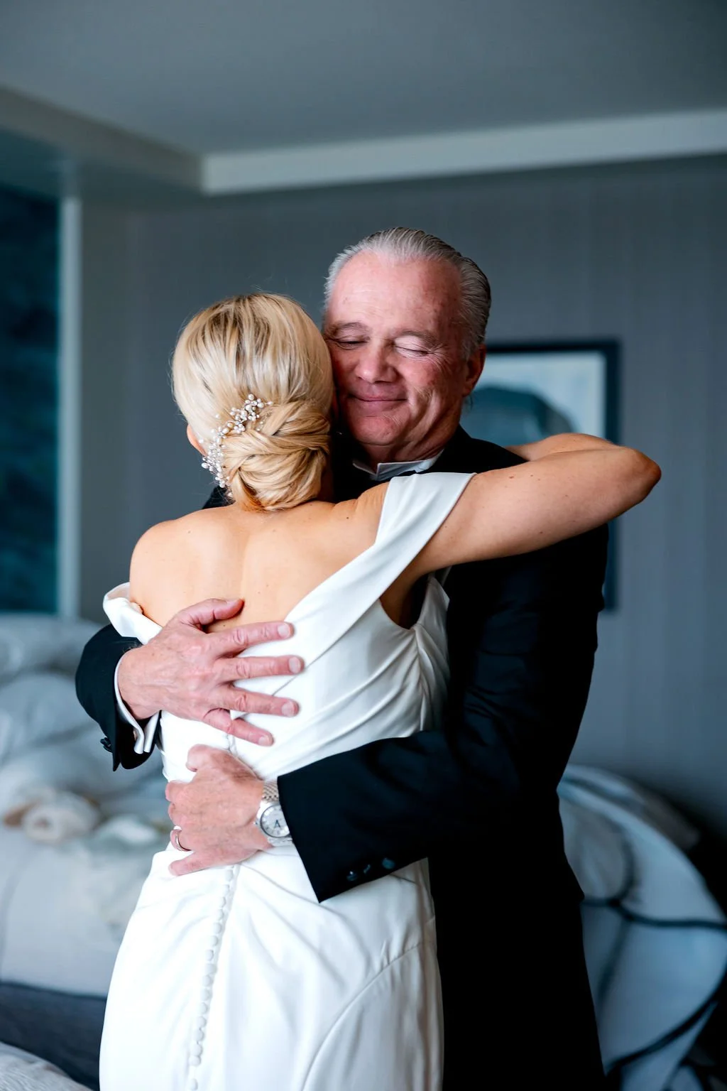 A bride and an elderly man share a warm hug in an indoor setting, with the bride's back visible and the man's eyes closed, both smiling.