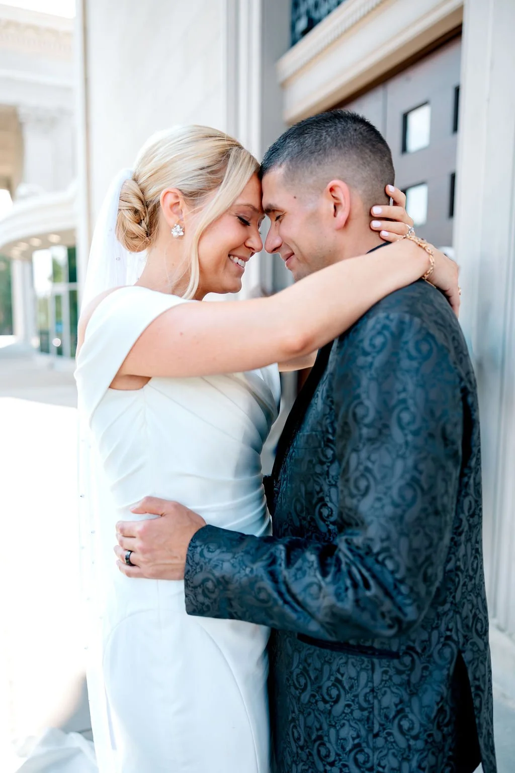 A bride and groom embrace, with foreheads touching, smiling and eyes closed outside a building.