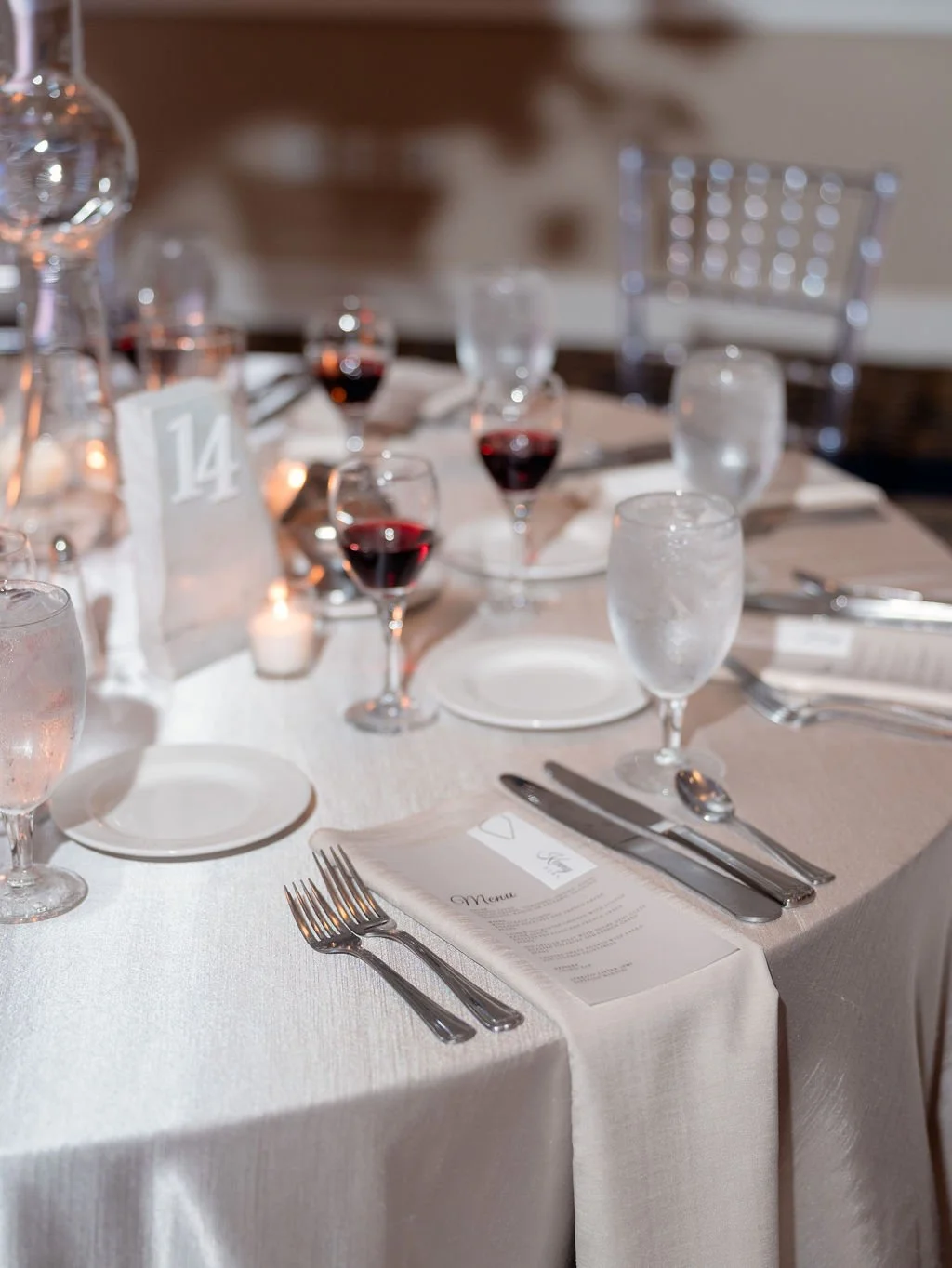 A formal dining table set with white tablecloth, water glasses, wine glasses filled with red wine, silverware, a menu card, and a table number sign with the number 14. Candles and chairs are also visible in the background.