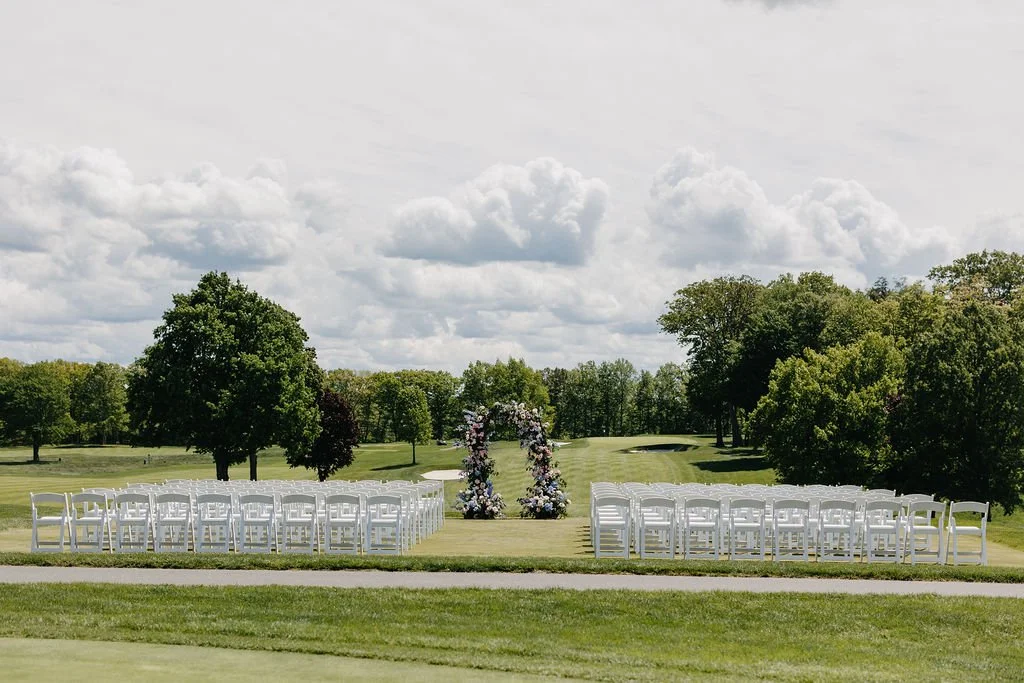 Outdoor wedding ceremony setup on a golf course with rows of white chairs facing a floral arch, surrounded by trees under a partly cloudy sky.