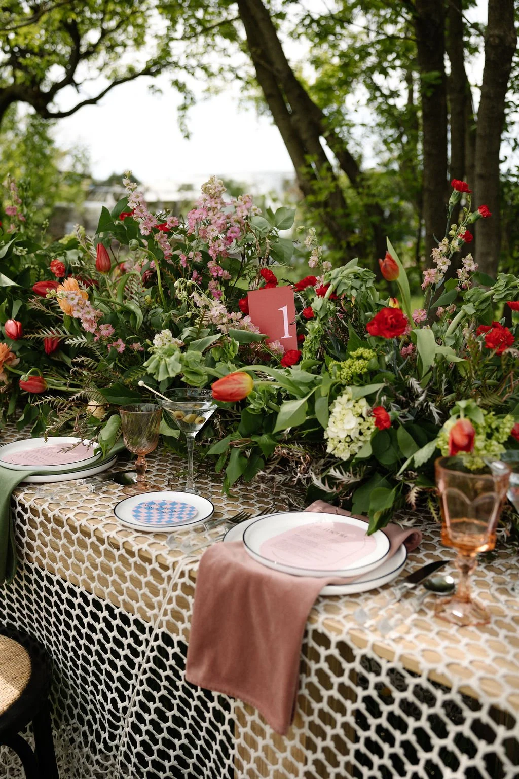 A beautifully set outdoor dining table decorated with an abundant arrangement of pink, red, and white flowers and greenery, featuring plates, glasses, and pink-toned napkins, with a pink table number card displaying the number one.
