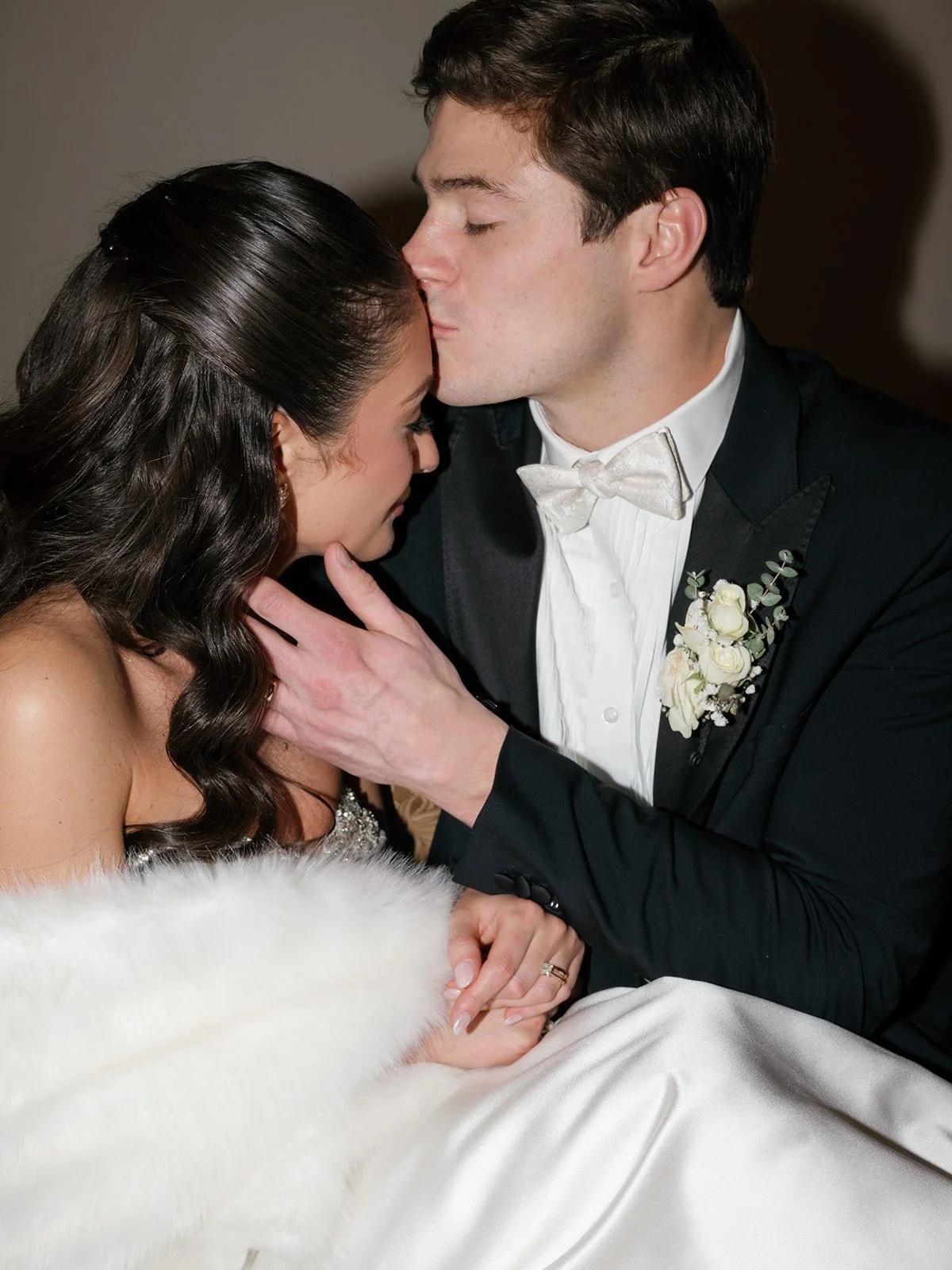 A groom kissing a bride on her forehead during their wedding ceremony, with the bride wearing a strapless dress and the groom in a black tuxedo with a boutonniere.