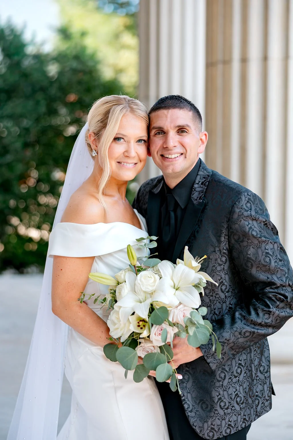 Bride and groom smiling while holding a bouquet of white lilies and pink roses outside near a columned building.