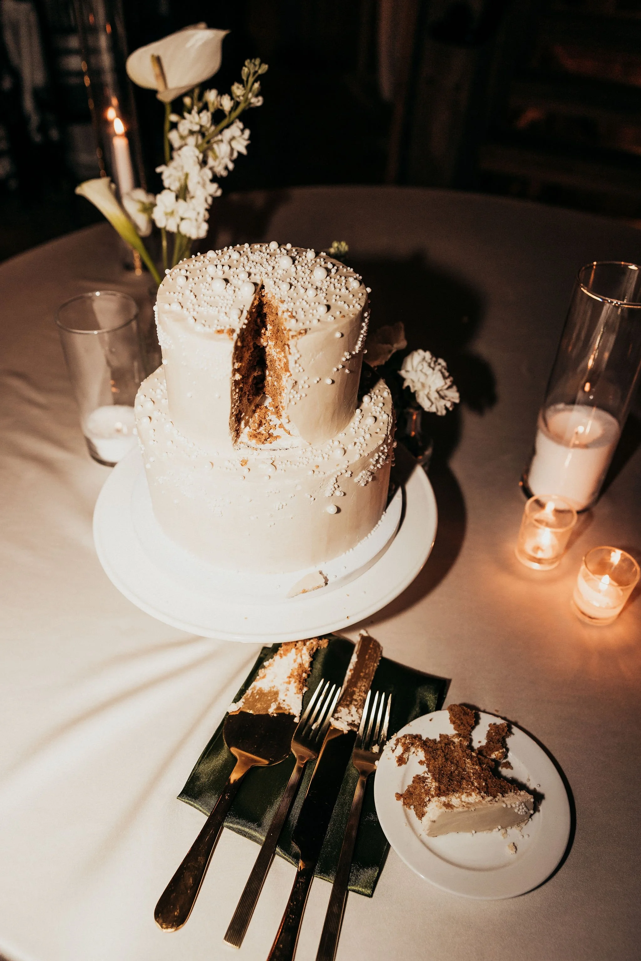 A two-layer vanilla cake with white icing and pearl decorations on top, partially sliced to reveal brown cake inside, on a white cake stand. Surrounding the cake are flowers in a vase, lit candles, a small plate with a slice of cake, and empty glasse