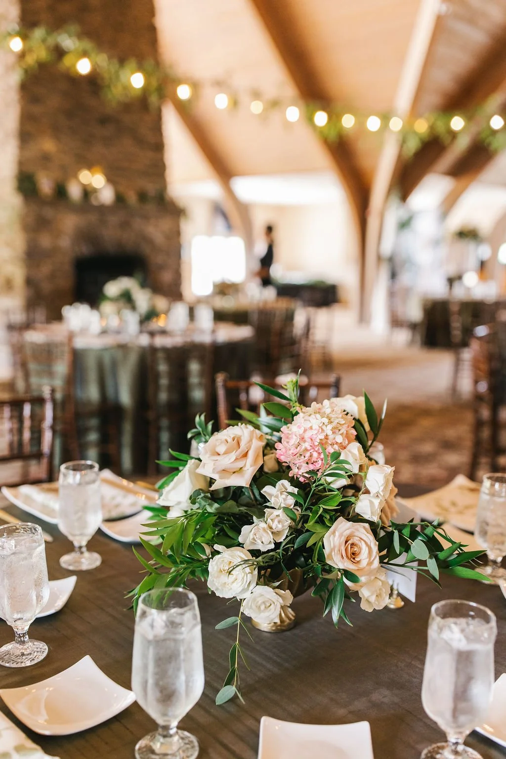 A wedding reception table with a floral centerpiece, water glasses, and white plates, set in a warmly decorated room with a stone fireplace and string lights.